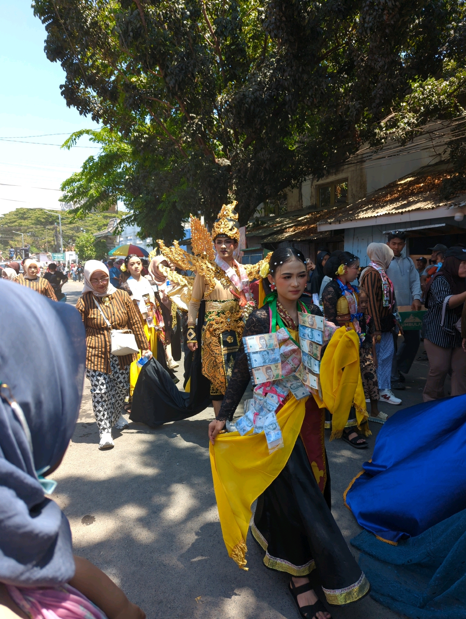 A procession is occurring on a paved street lined with buildings, under a bright sky and trees. Several people are participating, dressed in traditional clothing. In the foreground, a young woman in a black dress with a yellow sash adorned with banknotes walks forward, looking to her right. Behind her, a person in a golden costume with an elaborate headdress and wing-like embellishments is visible. To the left, an older woman in a striped brown and white outfit with a headscarf and a white purse walks ahead, holding a black cloth. Other participants in colorful attire are also present, with spectators on the side of the street. The environment suggests a cultural event or parade in a city, likely Pasuruan, Indonesia, given the context.