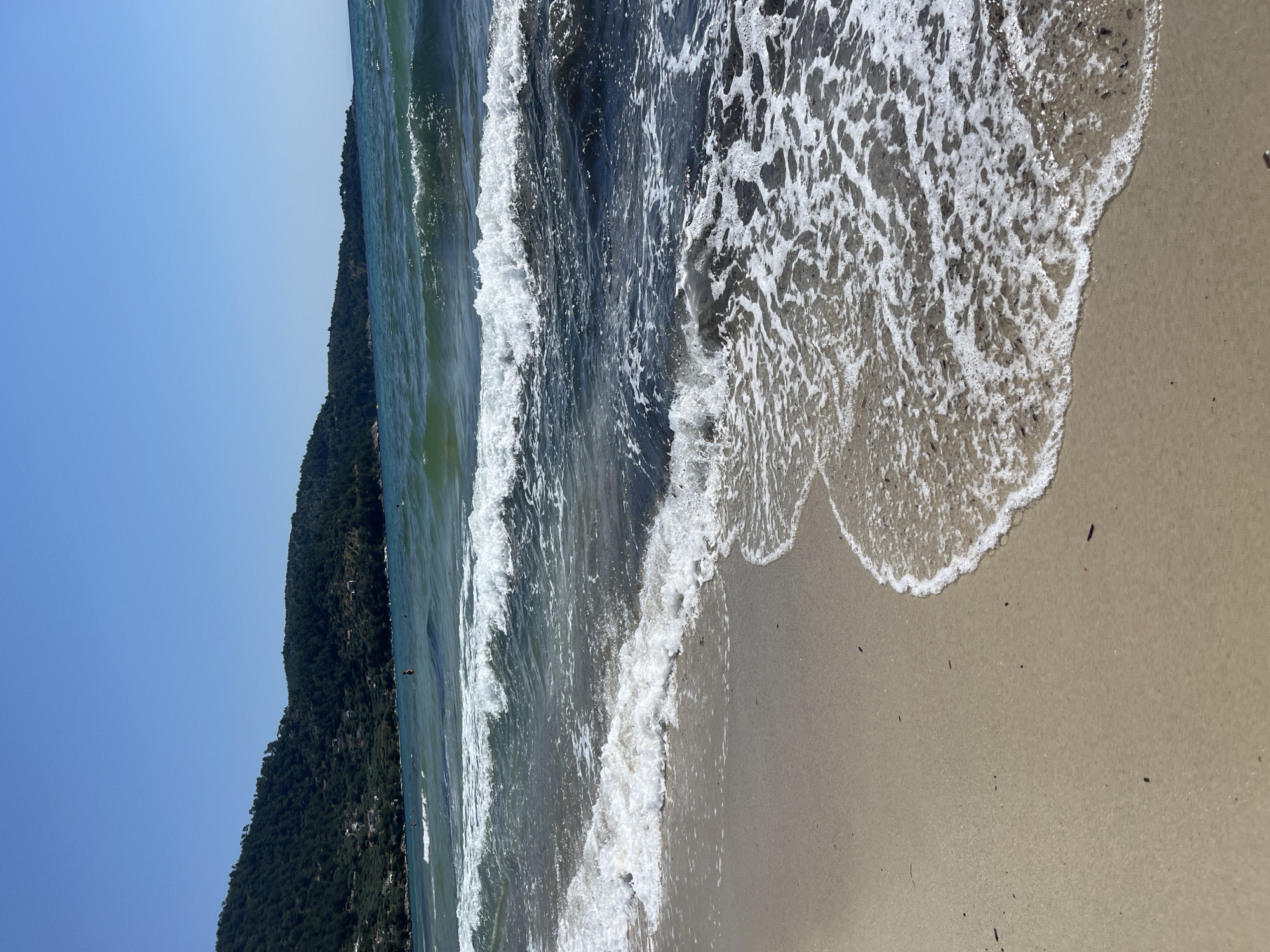 The image depicts a beach scene under a clear blue sky. In the foreground, a sandy beach is visible, with small pieces of debris scattered across it. Gentle waves are rolling onto the shore, creating white foam and ripples in the sand. The water is a mix of deep blue and green hues, with sunlight reflecting off its surface. In the middle ground, more waves are breaking. In the background, a densely forested hillside slopes down towards the sea. Small buildings are scattered amongst the trees on the hillside. The horizon line is sharp where the sea meets the sky. There are a few indistinct figures in the water, appearing to be swimming or wading. The scene is calm and tranquil, conveying a sense of a warm, sunny day at the beach.