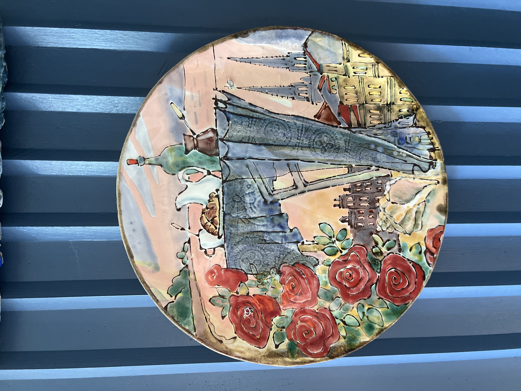 A circular, hand-painted ceramic plate depicts a whimsical scene with a breakfast table set against a backdrop of European city architecture. The plate is hung on a vertical, ribbed blue wall.
The foreground on the left side of the plate features a cluster of vibrant red roses with green leaves and stems. To the right of the roses, a table is draped with a dark blue tablecloth adorned with a delicate pattern. On the table, there is a white pitcher, two croissants, and a coffee pot.
Behind the table, a stylized depiction of a city skyline is visible. This includes buildings with red roofs and windows, and prominent church spires that suggest a Gothic architectural style. The background sky is a soft pink and orange, transitioning into lighter shades towards the top.
The plate itself has a golden-brown rim. A small metal hook is visible on the plate, near the top right, indicating it is hung on the wall. The blue wall has a matte finish and vertical grooves, creating a textured background. There are no people or animals present in the scene. The overall impression is a romanticized, artistic representation of a European breakfast setting.