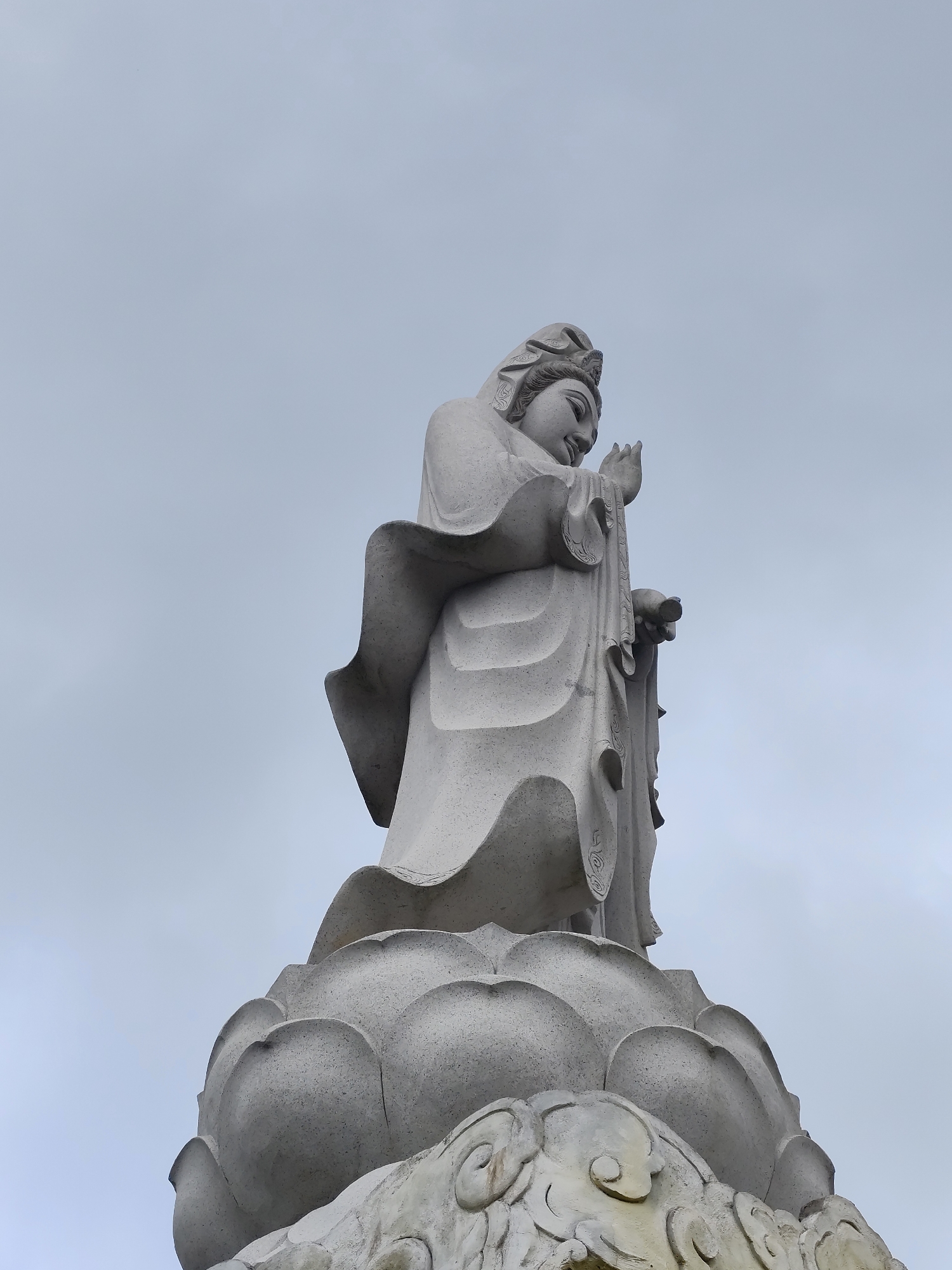 A low-angle, long shot captures a weathered statue of a robed figure, likely female, atop a lotus pedestal against a backdrop of a cloudy gray sky. The statue's features are gently aged, and it stands in the city of Cam Ly - Đà Lạt, Vietnam. The figure's hands are positioned near each other, possibly in a gesture of blessing or offering. The lotus pedestal is detailed with petal shapes and cloud-like patterns at its base. The overall composition emphasizes the statue's height and serene presence against the overcast sky.