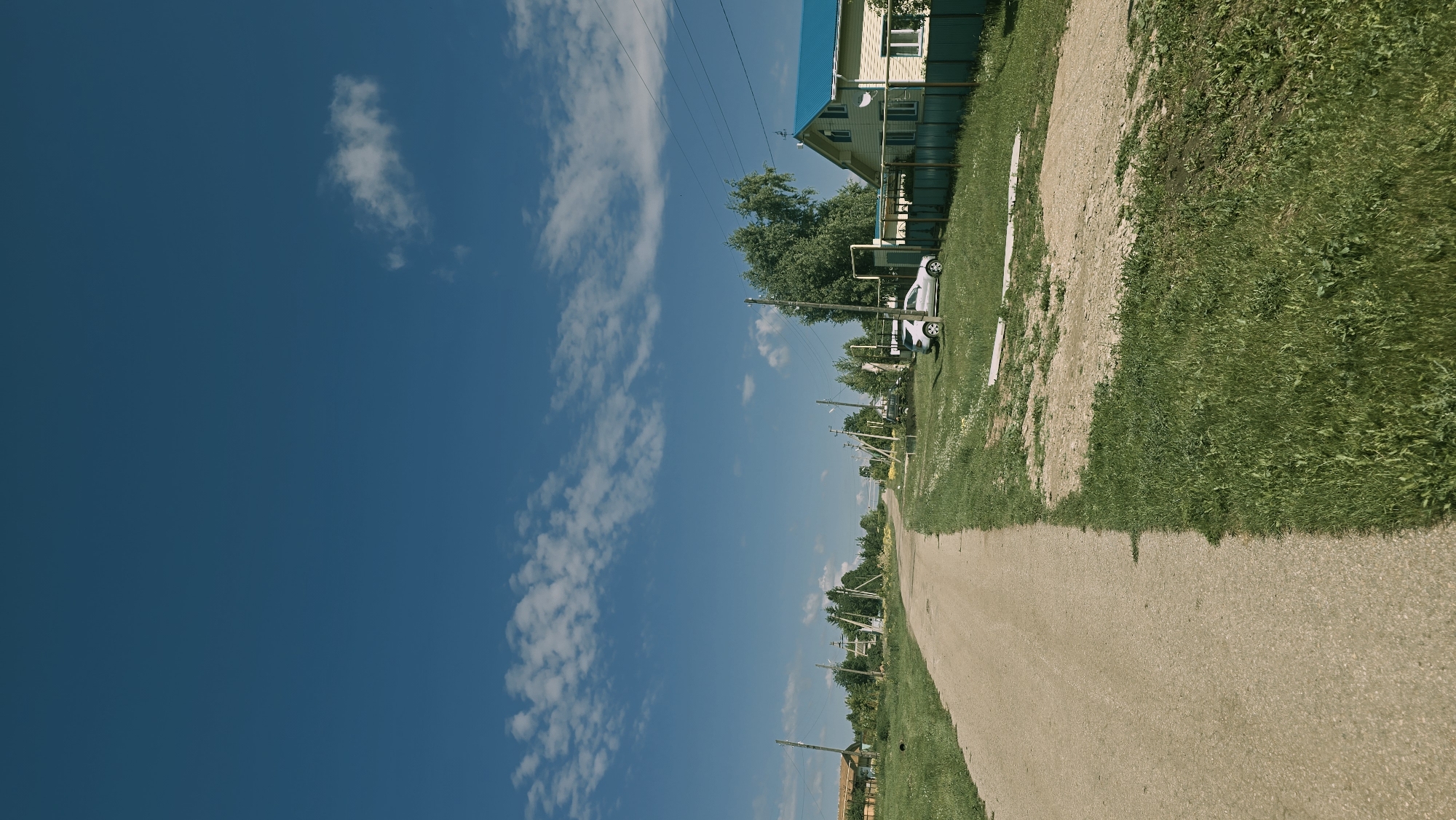 The image depicts a rural scene on a sunny day in Kzyl-Chishma, Russia. A dirt road runs from the foreground into the distance, bordered by green grass and some sparse vegetation. Several utility poles with wires are spaced along the side of the road. In the middle ground, a light-colored car is parked on the right side of the road, near a house with a yellow facade and a bright blue roof. A green fence surrounds the property. Further in the distance, more houses and trees are visible, suggesting a small village or settlement. The sky is a clear, bright blue with a few scattered white clouds. The overall impression is one of a quiet, summer day in a rural Russian locality.