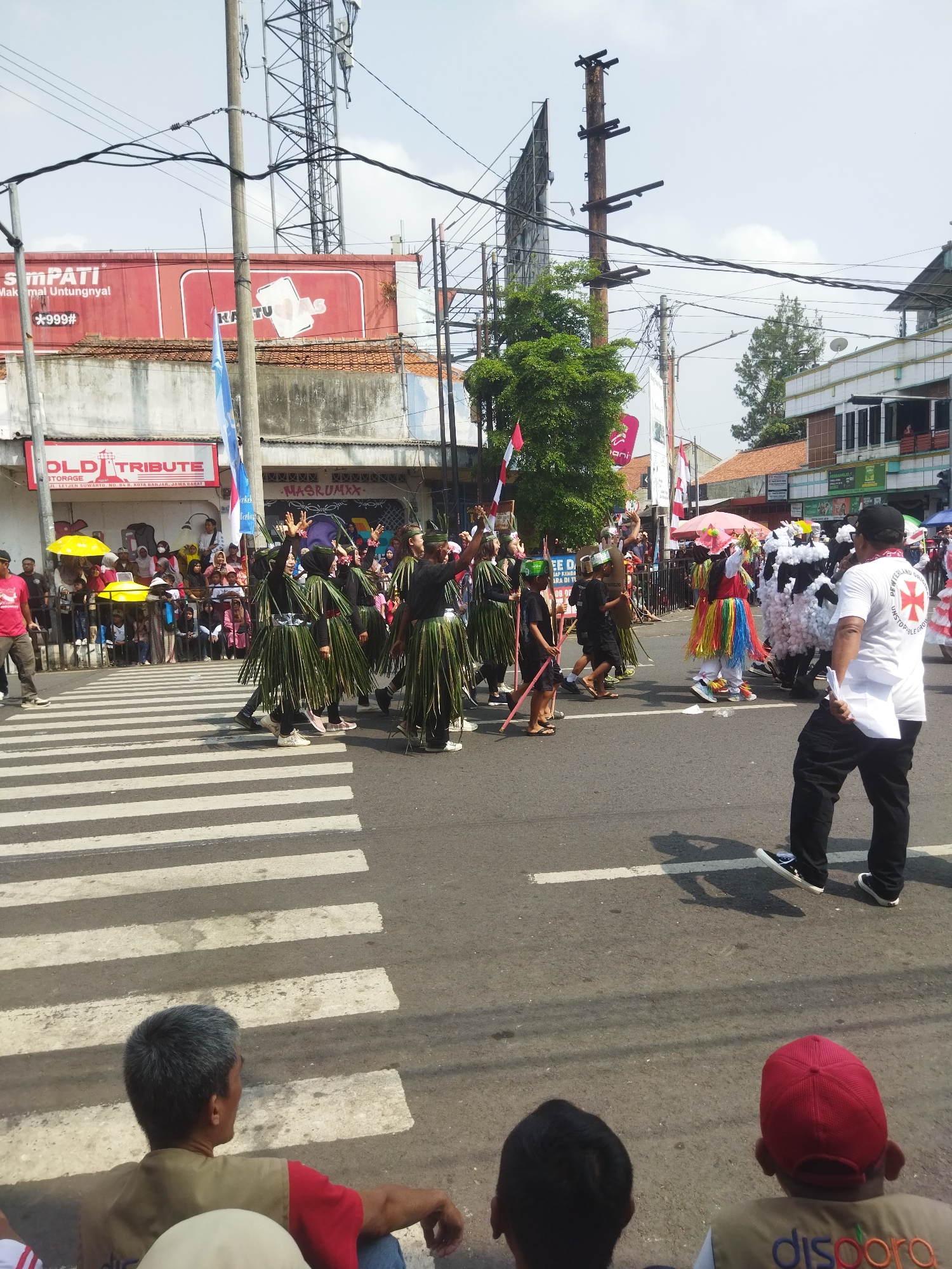 The image captures a street parade in Banjar, Indonesia, likely on Jalan Letjen Suwarto, indicated by signage. Several individuals, dressed in unique costumes resembling grass skirts, are crossing a pedestrian crosswalk. Some are wearing green, leafy garments, while others are adorned with colorful, fringed outfits or white fluffy costumes. One person in the foreground on the right wears a white t-shirt with a red cross emblem and the text "PETERBAND UNSTOPPABLE". The parade appears to be taking place on a sunny day with a clear sky. Spectators are visible on the left side of the road, behind a barrier, watching the procession. Buildings line the street, with some featuring advertisements, including one for "SIMPATI" and "OLD TRIBUTE". Utility poles and overhead wires crisscross the sky, suggesting an urban setting. The overall scene depicts a festive community event in progress.