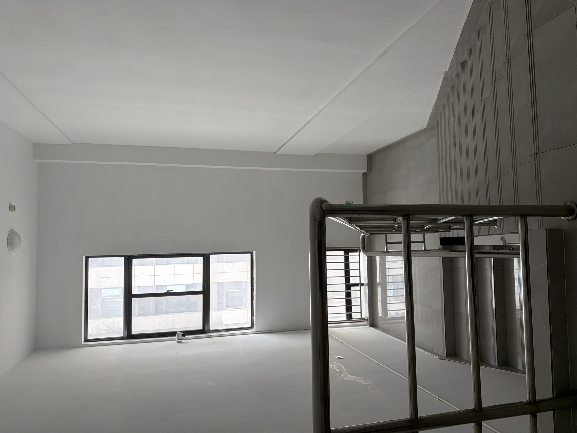 The image depicts the interior of a building, likely a stairwell, in Jiangning District, China. The space is mostly white and gray. Two sets of windows are visible, offering a view of other buildings outside, indicating an urban environment. The focus is on the metal railings and staircase, suggesting a modern design. The scene is sterile and lacks any human presence or personal items. The environment is clean and appears to be newly constructed or renovated.