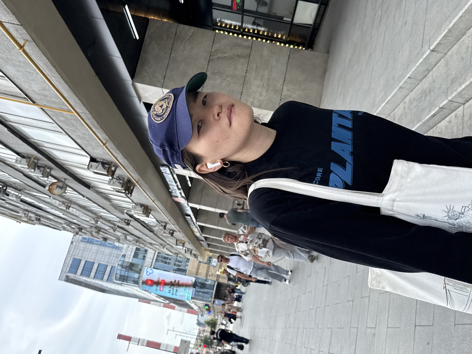 A woman wearing a dark long-sleeved shirt, a blue baseball cap, and white wireless earbuds is walking on a paved sidewalk in Moscow, Russia. She is carrying a white tote bag over her left shoulder. The woman is looking towards the camera with a neutral expression. In the background, there are buildings with numerous windows and air conditioning units, as well as other pedestrians walking on the sidewalk. The sky is overcast. The scene appears to be a commercial or business district of the city.