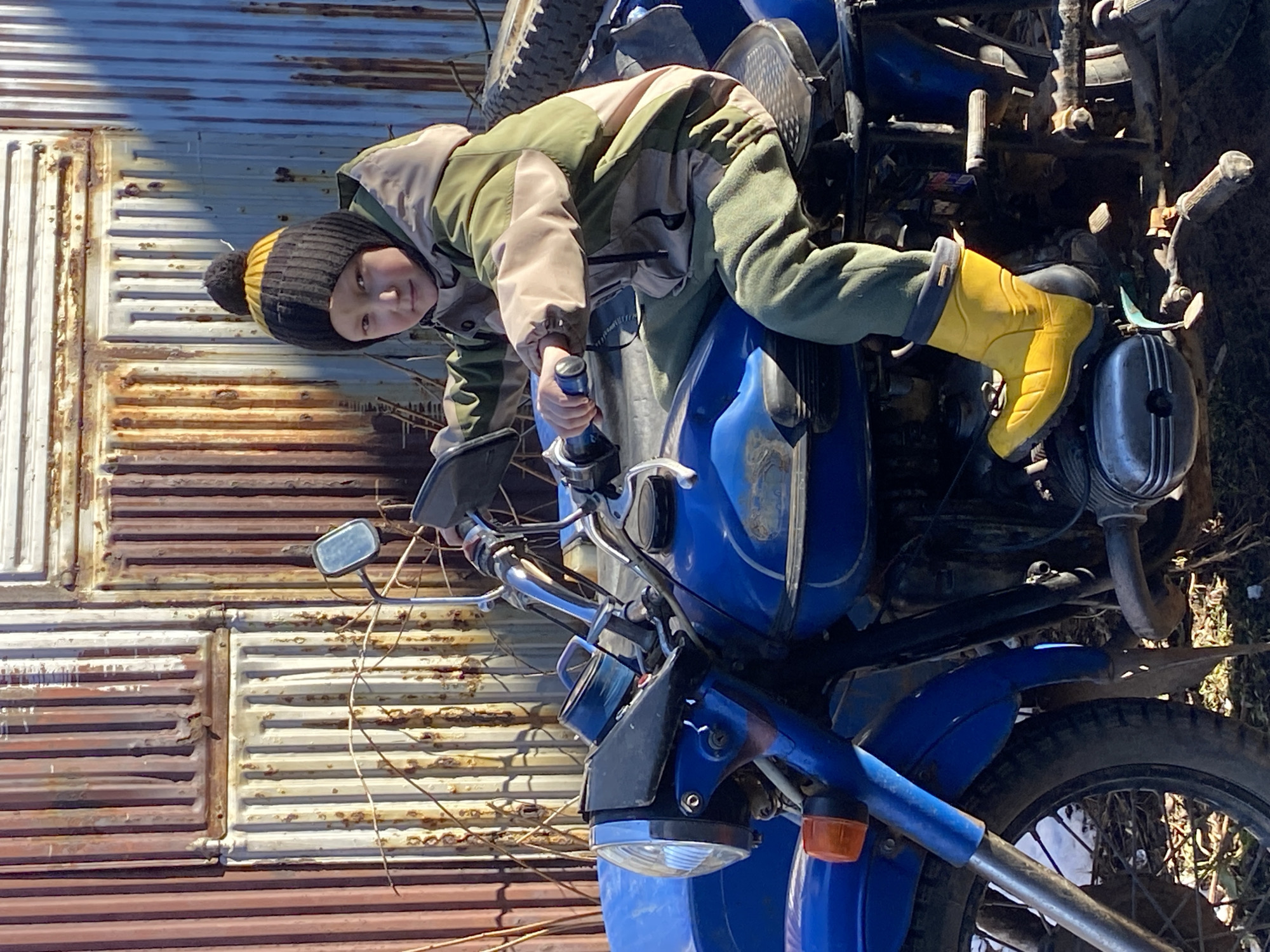 A young child, wearing a khaki and tan jacket, green pants, a black and yellow beanie with a pom-pom, and yellow boots, is seated on a blue motorcycle. The motorcycle is positioned in front of a corrugated metal wall or shed. The location is possibly Noviy, Russia, based on the instruction, but this cannot be verified from the image alone. The background includes some sparse vegetation and debris. The motorcycle appears old or in disrepair. The child is holding the motorcycle's handlebars.