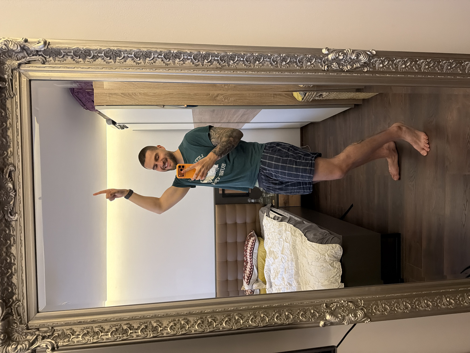 In a residential interior, a man is taking a mirror selfie. He is wearing a green t-shirt and plaid shorts, and has tattoos on his arm. He is smiling and pointing upwards with his right index finger. Behind him, a bed is visible, along with a wardrobe and a mirror with an ornate frame. The flooring is dark wood. The image is taken from the perspective of looking into the mirror, showing the man and his reflection, as well as the room behind him. There is no indication of the city of Sofia, Bulgaria.