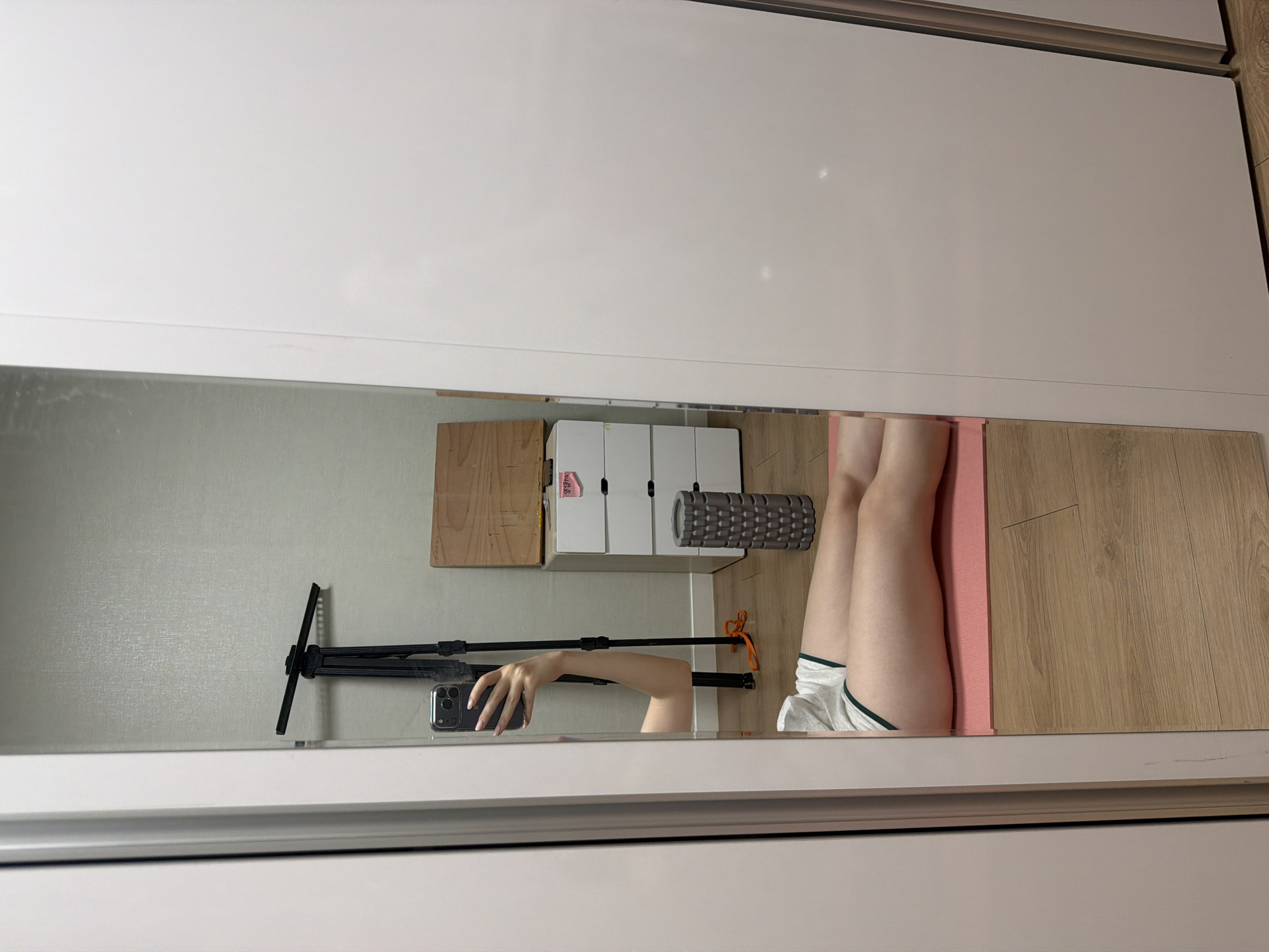 A person is sitting on a pink yoga mat on a wooden floor, reflected in a large mirror. They are wearing white shorts with green trim and a white tank top. Their legs are extended in front of them. In the reflection, their arm is raised, holding a smartphone, and their face is visible, looking at the phone. Behind the person in the reflection, there is a black tripod, a grey foam roller, a white chest of drawers with pink labels, and a wooden panel. The background is a wall with a textured pattern. The image appears to be taken indoors, likely in a home or gym setting in Dongducheon-si, South Korea.
