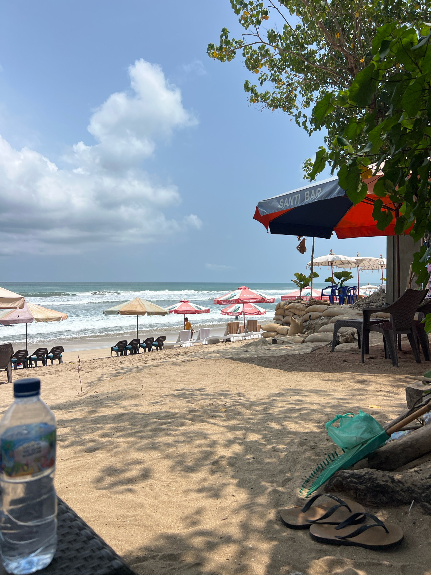 A beach scene in Kuta, Indonesia, shows a sandy shore with lounge chairs and umbrellas. In the foreground, a plastic water bottle sits on a woven surface, with a pair of flip-flops, a rake, and a green plastic bag on the sand. Behind this, the beach stretches towards the ocean, where waves are breaking. Several beach umbrellas are set up, with lounge chairs arranged beneath some of them. A bar area is visible in the mid-ground, featuring a large umbrella with "SANTI BAR" printed on it, and sandbags forming a low wall. In the distance, more umbrellas and lounge chairs dot the beach, and a person in a yellow shirt stands near the water. The sky is partly cloudy.