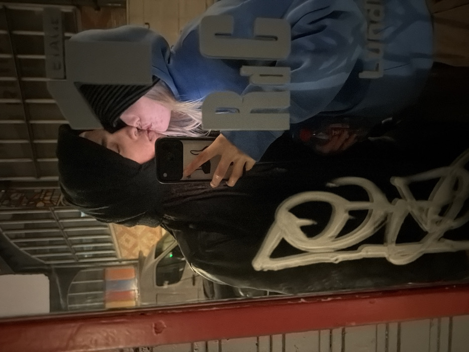 A mirror reflects two people kissing. The person on the right wears a light blue hoodie with white lettering. The person on the left wears a dark hoodie and a striped hat. One of the people holds a phone with a clear case and a black cat design. A car and some barrels are visible behind the couple in the mirror's reflection. A white graffiti design is at the bottom of the frame. The mirror has the word "Luride" visible in the reflection.
