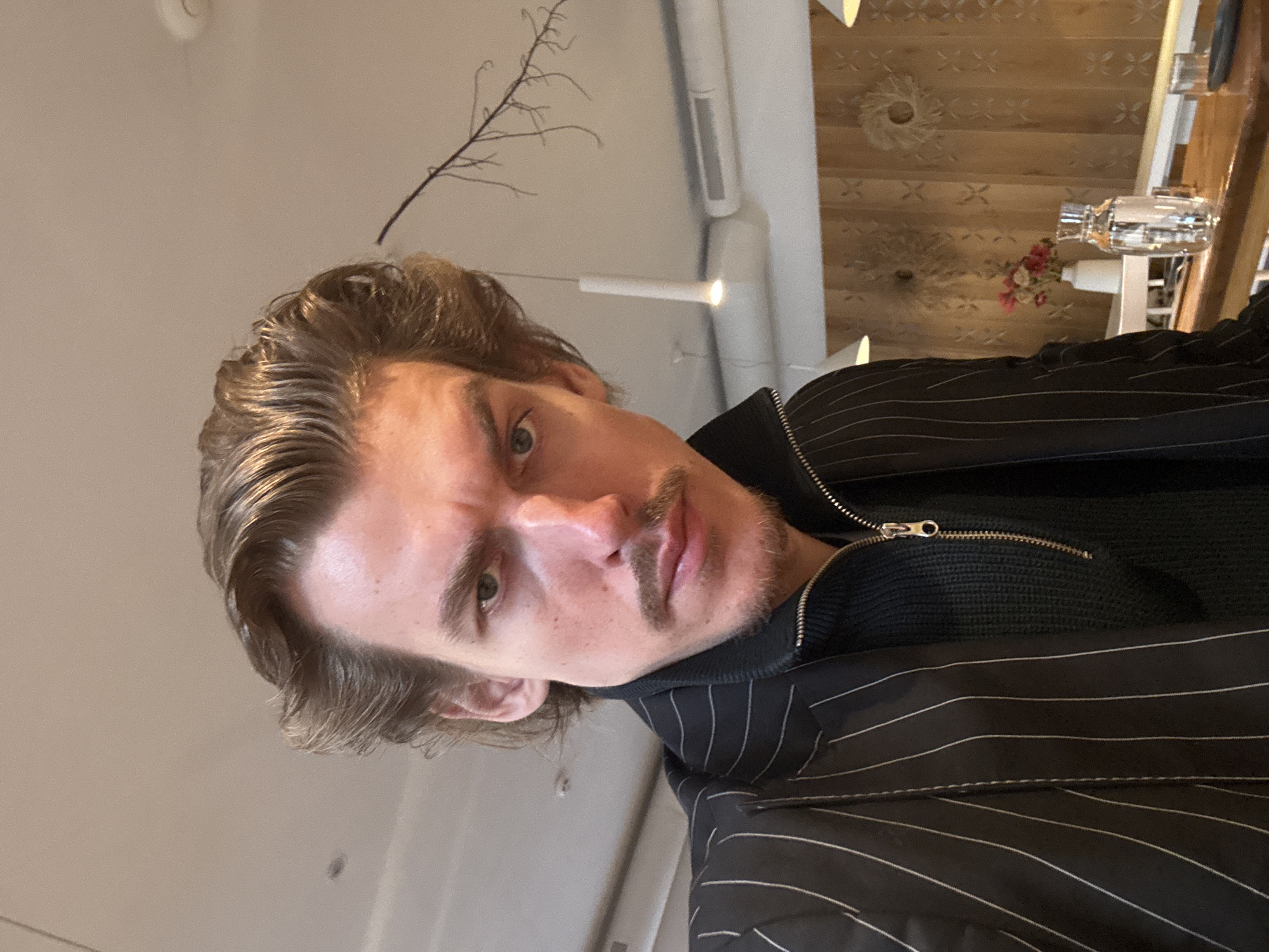 A selfie of a person with shoulder-length brown hair, a mustache, and green eyes. The person is wearing a dark-colored, horizontally striped jacket over a dark sweater. The background includes a ceiling with decorative wooden panels, white support structures, and a light fixture. The exact location in Kyiv, Ukraine cannot be determined without additional information.