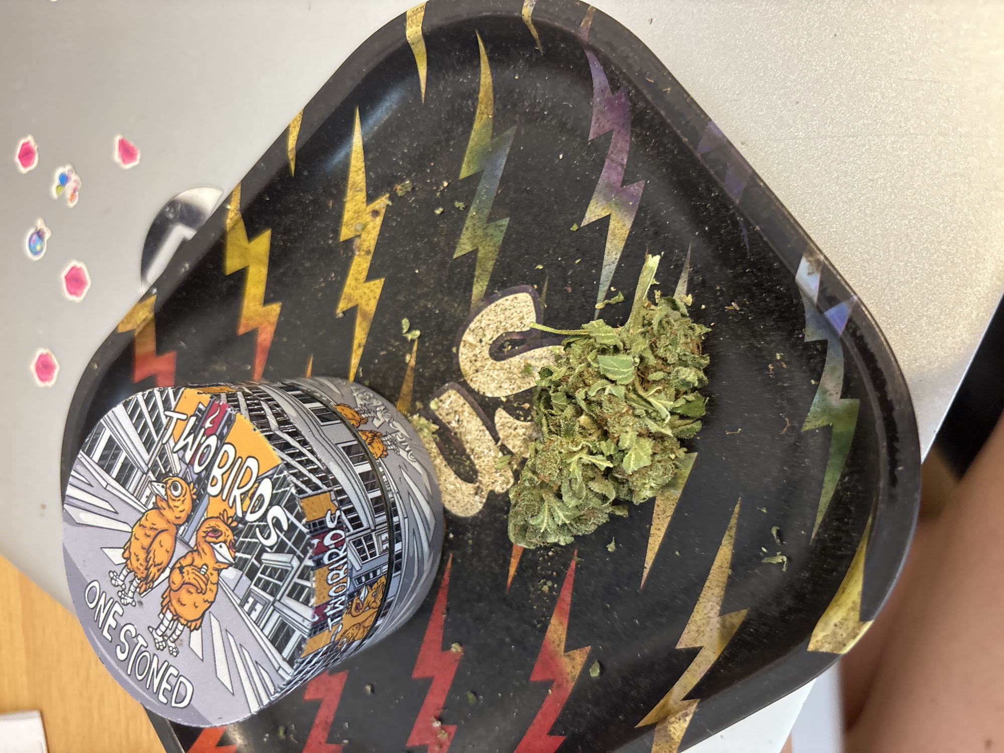 A top-down, eye-level view shows a "Two Birds One Stoned" rolling tray, cannabis, and related paraphernalia on a light surface. The rolling tray is black with colorful lightning bolt designs and some cannabis residue. A grinder with the same "Two Birds One Stoned" design sits on the tray. To the right of the grinder, there is a clump of green cannabis. Stickers are visible on the light surface near the tray. The specific location in Buenos Aires, Argentina, cannot be determined from the image. No people are fully visible, but a small portion of a leg is present at the edge of the picture.