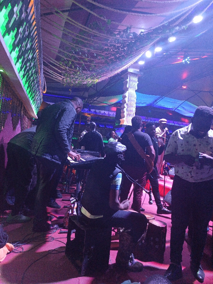 Inside a dimly lit venue, a band performs on stage. A keyboard player, clad in a black jacket, stands to the left, his hands poised over the instrument. A seated drummer plays a small, wooden drum to his right. Other musicians, including a guitarist, are visible behind them. The stage is bathed in purple light, with greenery hanging from the ceiling. Audience members stand around, engrossed in the music.