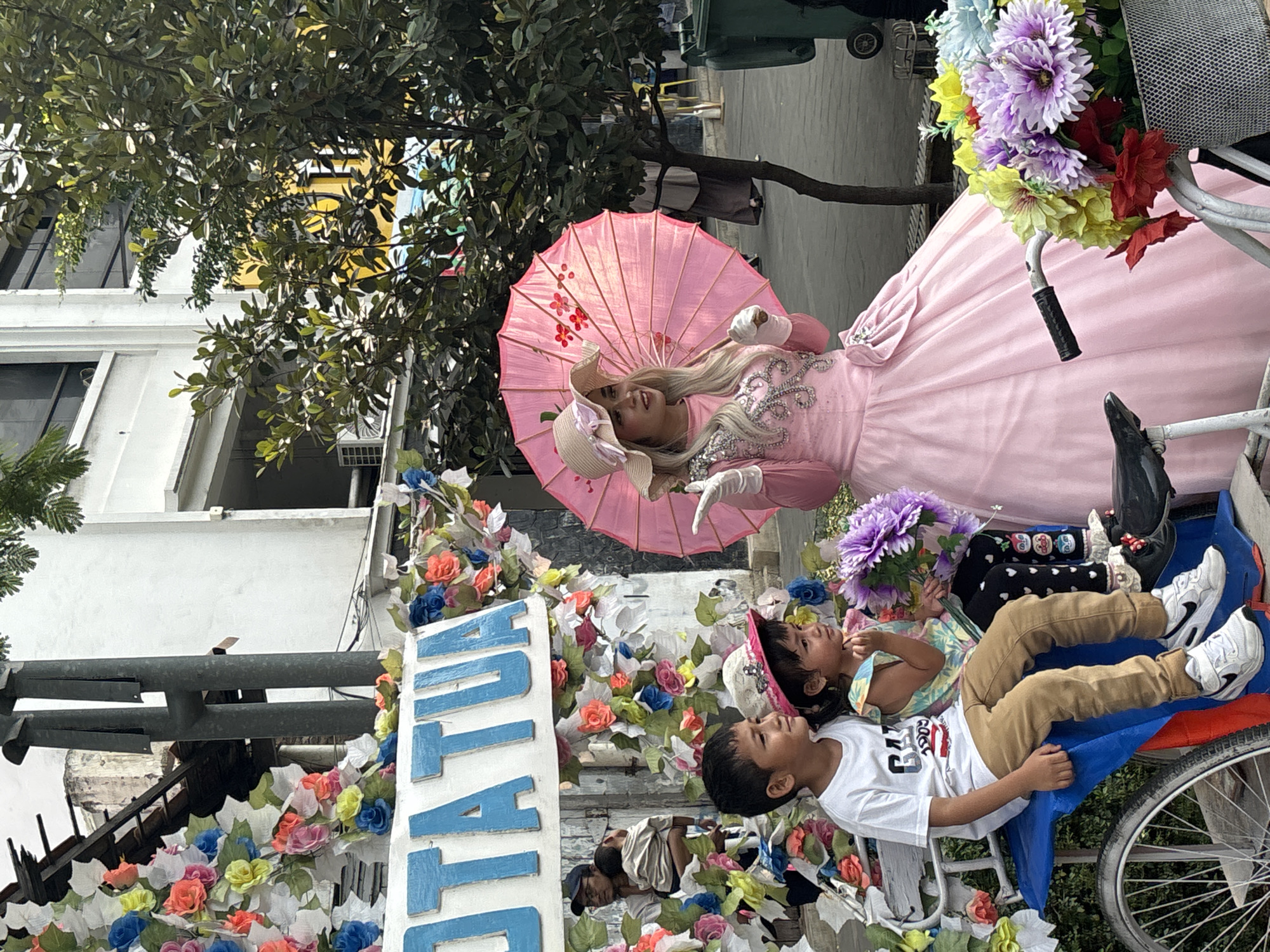 In the Kota Tua area of West Jakarta, Indonesia, a woman in a pink ruffled dress and a wide-brimmed hat stands holding a pink umbrella with floral accents. She wears white gloves and has blonde hair. In front of her, two children are seated on a blue structure that appears to be part of a pedicab or bicycle. A young boy in a white t-shirt and brown pants sits on the left, and a young girl in a colorful top and patterned leggings sits to his right. Both children are looking to the right. The scene is decorated with colorful artificial flowers, and a bicycle with a basket of flowers is visible in the foreground. In the background, a building with windows and trees can be seen. A man carrying a child is also visible in the background to the left.