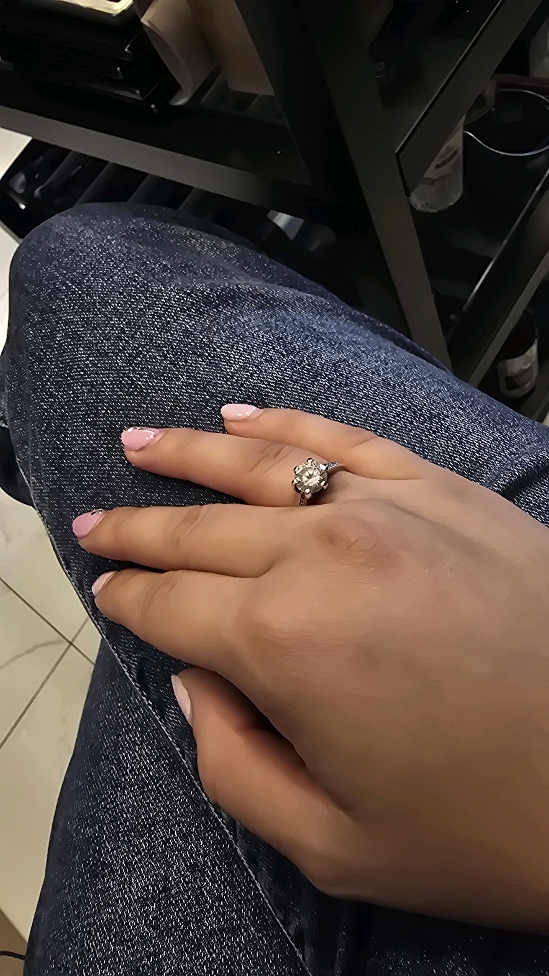 A close-up, high-angle shot focuses on a person's hand, adorned with a ring, resting on their lap covered by dark blue denim jeans. The ring features a prominent, clear stone set within a delicate, floral-shaped metal band. The fingernails are painted a soft pink with French tips. The background is blurred, showing dark furniture with shelves. The scene appears to be indoors. The location is Pazardzhik, Bulgaria.