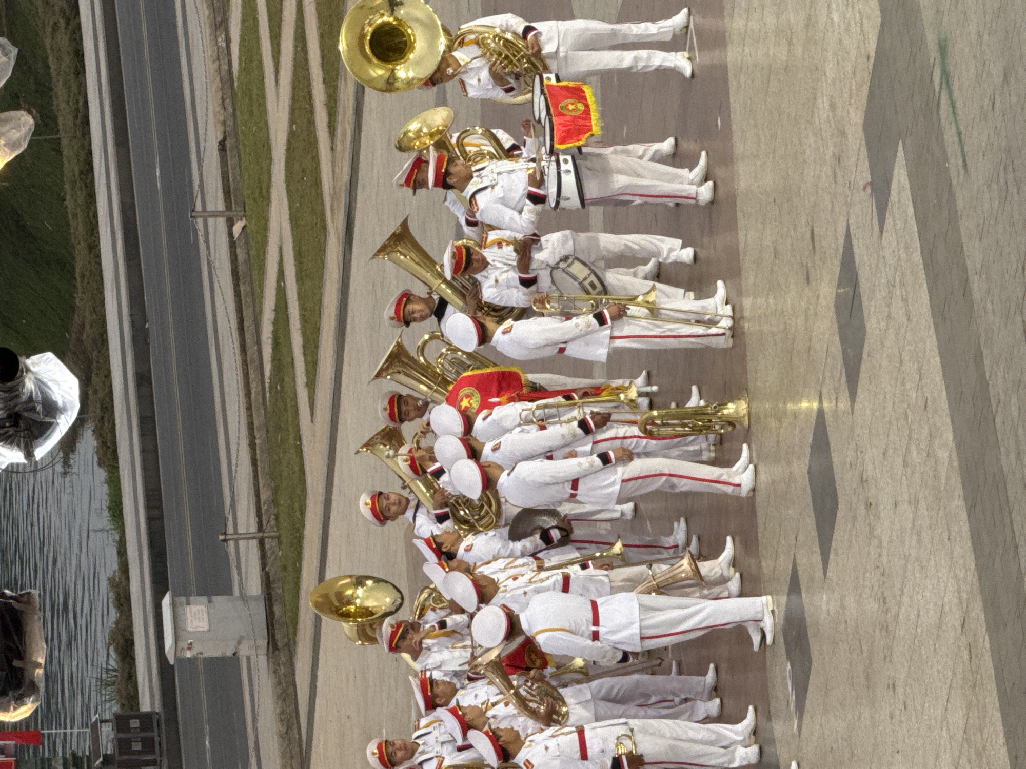 The image depicts a marching band in uniform, standing on a paved plaza. The band members are predominantly male, wearing white uniforms with red trim and white caps. They are holding various brass instruments, including tubas, trombones, and trumpets. Two individuals in the foreground are holding drums and a red banner with a yellow emblem. The background shows a body of water with a distant road and some greenery. The scene is likely a public event or ceremony. The presence of the band and the formal attire suggests a parade or performance. The paved plaza indicates an urban setting, and the water in the background could be a lake or river. The overall atmosphere appears to be organized and ceremonial.