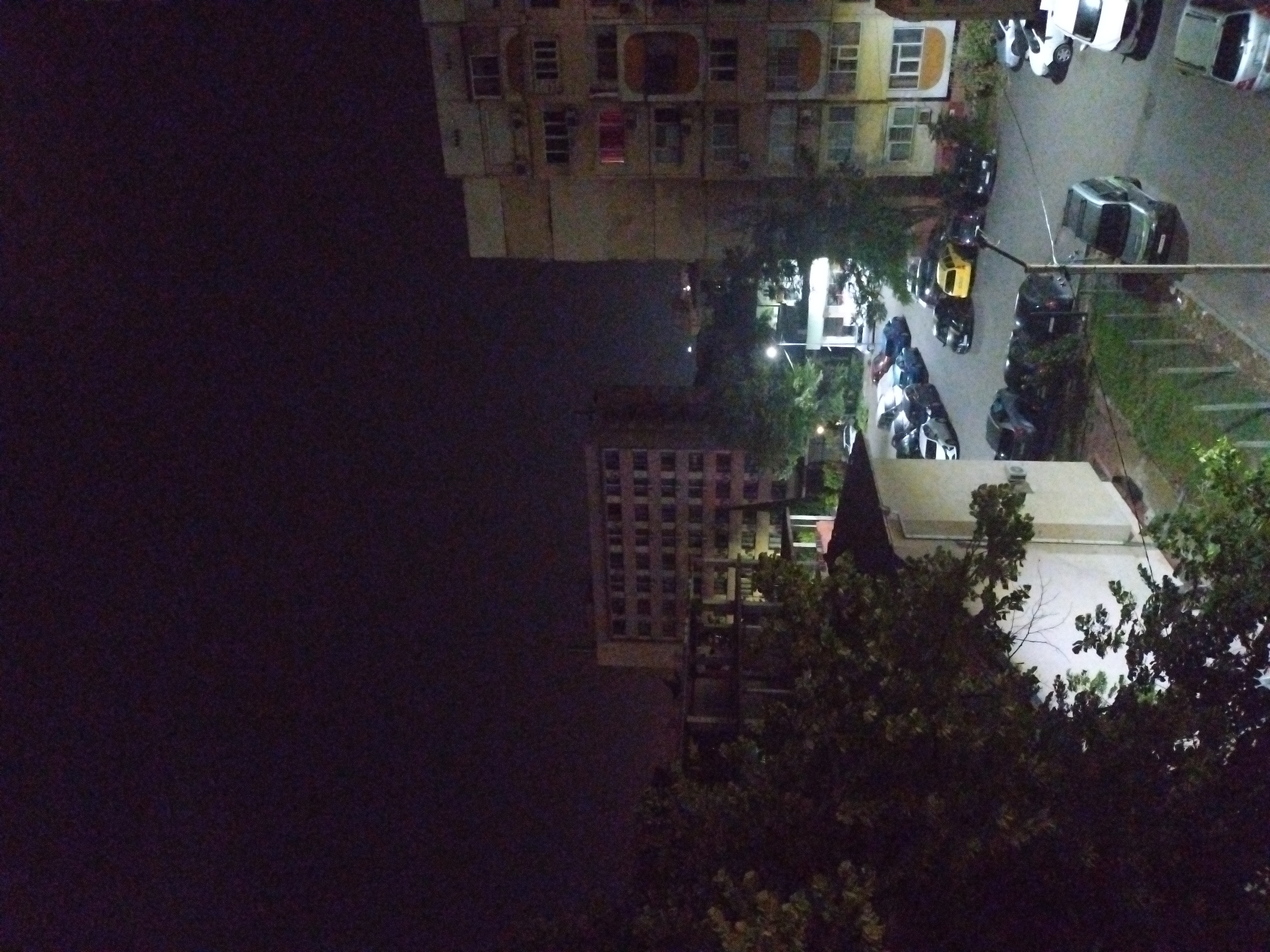 The image depicts a nighttime residential urban environment in Stara Zagora, Bulgaria, viewed from an elevated perspective. The scene is dominated by multi-story buildings and numerous parked vehicles, illuminated by streetlights and interior lights from windows.
In the foreground, dense, dark foliage from trees and bushes occupies the lower left portion of the frame, partially obscuring a light-colored, low-rise building. A prominent streetlight is visible in the mid-left, casting light onto a paved parking area.
Two large multi-story residential buildings are central to the composition. On the left, a tall, uniformly structured building with multiple illuminated windows stands. To the right, another multi-story apartment block features a beige facade with varying window designs, balconies, and air conditioning units. Several windows in this building are lit, with one window on an upper-middle floor exhibiting a distinct red light.
Between and around these buildings, multiple parking lots and street sections are densely packed with parked vehicles. These include various models such as sedans, hatchbacks, SUVs, and a minivan, in colors including white, silver, black, blue, and a distinct yellow car in the mid-left parking area. A silver minivan/SUV is prominently parked in the lower right foreground. Paved surfaces for parking and roads are visible, alongside small patches of grass and fencing.
The sky is dark, indicating night, with visible image noise consistent with low-light photography. No individuals are visible, and no active events or interactions are depicted; the scene appears static. Distant lights suggest the continuation of the urban area in the background.