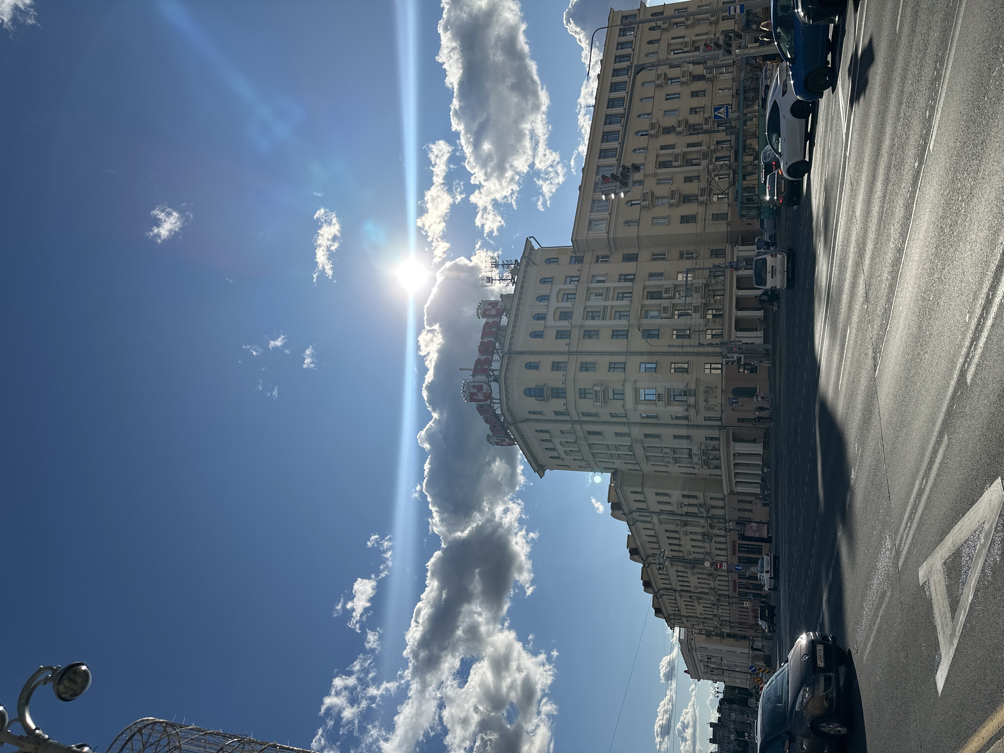 In Minsk, Belarus, on a bright, sunny day with scattered clouds, a street scene unfolds. A modern, multi-story building with a distinctive red sign on its roof stands prominently. The building has a light yellow facade and numerous windows, with some apartments featuring balconies. The street in front is paved, with visible lane markings and a pedestrian crossing. Several cars are present, including a dark sedan in the foreground and other vehicles of various colors in the background. People are seen walking on the sidewalk and crossing the street. A street lamp with a decorative design is visible on the left side of the frame. The sun is shining brightly, creating lens flares and casting shadows across the road. The overall impression is of a typical urban environment in daylight.
