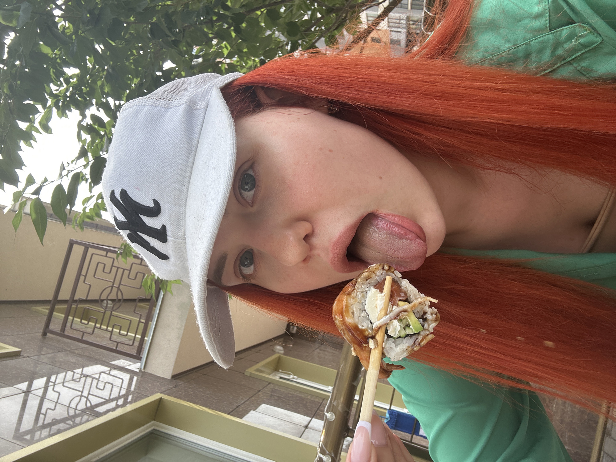 In Lymanka, Ukraine, a close-up shot features a young woman with vibrant red hair and a white baseball cap with a black emblem. She is wearing a green top and is in the process of eating a piece of sushi with chopsticks. Her tongue is extended, reaching towards the sushi. The background shows a building with an ornate balcony and green foliage from trees.