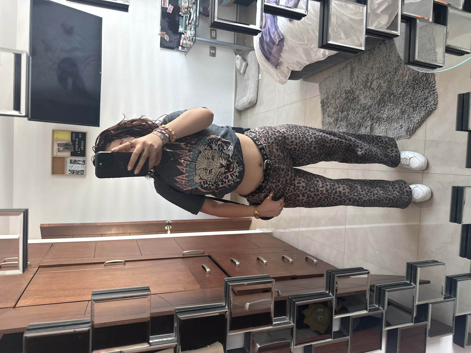 A person stands in front of a mirror, taking a photo of themselves with a mobile phone. They are wearing a graphic t-shirt, leopard-print pants, and white sneakers. The background includes a bed with bedding, a gray rug, a TV screen, wall decorations, and a dark wood dresser with mirror accents. The location appears to be a residential room in Santa Clara Ocoyucan, Mexico.