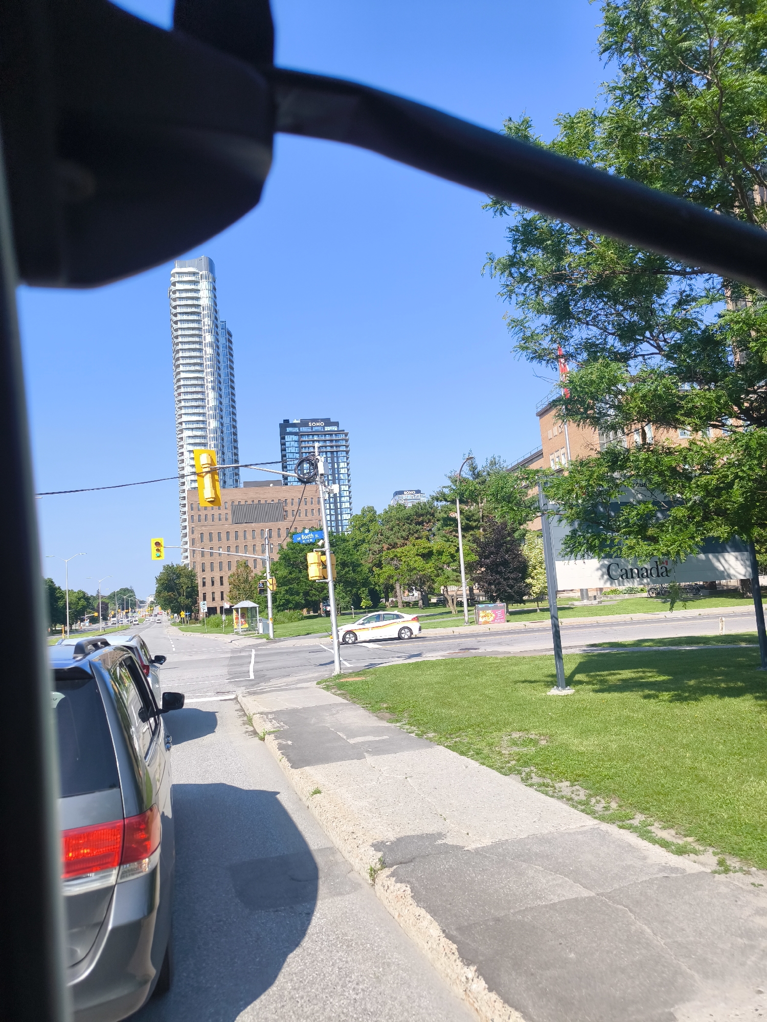 The image captures a street scene in Ottawa, Canada, likely during daylight hours under a clear blue sky. The perspective appears to be from inside a vehicle, with a dark, partially obscured frame in the foreground suggesting a window or part of the car's interior.
In the mid-ground, a multi-lane intersection is visible with traffic lights. Several cars are present on the road, with one dark-colored SUV prominently featured in the lower left, its red taillight illuminated. Across the intersection, a light-colored sedan is turning. Beyond the intersection, a bus stop shelter is situated on the right side of the road, adjacent to a manicured green lawn.
Dominating the background are several modern buildings. A tall, slender skyscraper with a white facade and numerous balconies stands out against the sky. Next to it, a darker, rectangular building displays "SOHO" in white lettering. Another brick building, lower in height, is situated between these two taller structures. On the far right, a large sign with "Canada" in prominent lettering is visible on the side of a building. Trees with lush green foliage are scattered throughout the scene, particularly on the right side of the image. The overall environment suggests an urban setting with a mix of residential and commercial architecture.