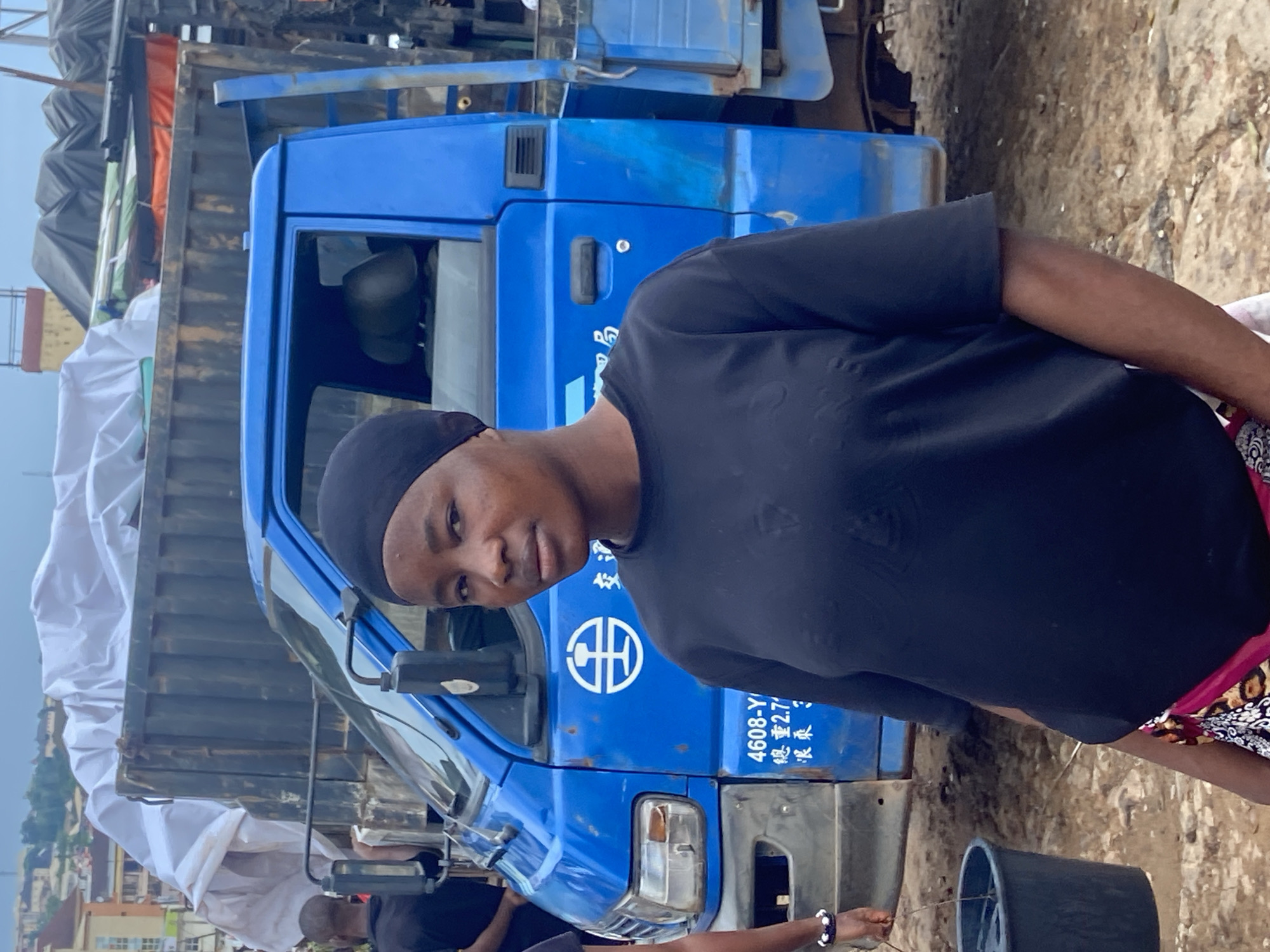 The media file captures a person standing next to a blue truck in Ogbunike, Nigeria. The person is wearing a black t-shirt and a black head covering. Their expression is neutral. The truck appears to be a flatbed truck, with some items covered by white tarps in the bed. There is also some cargo visible behind the truck, partially obscured by tarps of various colors, including orange and green.
The setting appears to be an outdoor area, possibly a roadside or a yard, with a dirt or gravel ground. In the background, there are buildings and some greenery, suggesting an urban or semi-urban environment. The sky is visible and appears overcast, indicating a cloudy day.
Visible text on the truck includes a logo with a circular design and some characters that appear to be Chinese. Below this, there is numerical text "4608-Y" and other characters. There is also text in what appears to be Chinese characters below that.
The image is oriented vertically, suggesting it may have been taken with a mobile phone. The lighting suggests it is daytime, but the overcast sky does not provide strong shadows. The overall impression is a candid snapshot of daily life.