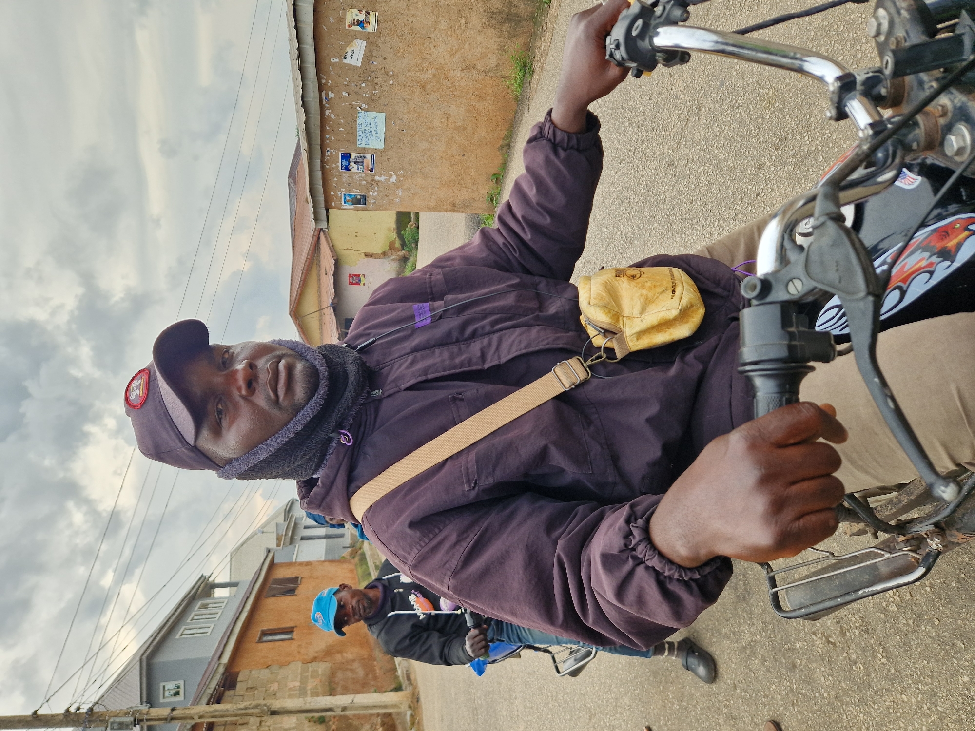 A man wearing a dark baseball cap, a dark purple hooded jacket, and a light brown crossbody bag is riding a motorcycle. He has a dark neck gaiter pulled up around his neck. Another man wearing a blue baseball cap and a black hoodie is riding on the back of the motorcycle. They are on a paved road in an urban area with buildings on either side of the street. The buildings appear to be a mix of residential and commercial. The sky is overcast. The image is taken from a low angle, looking up at the man riding the motorcycle.