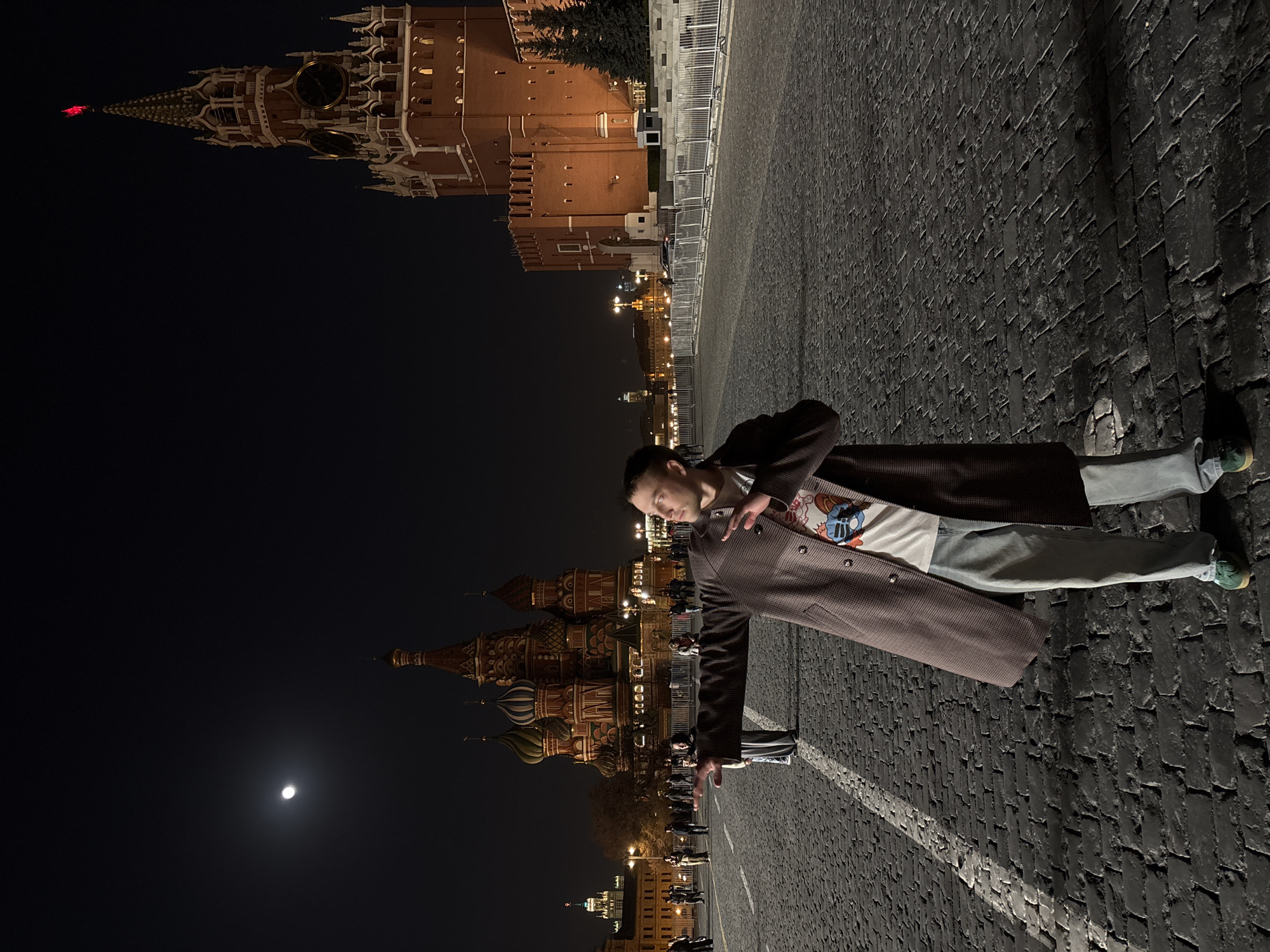 At night in Moscow, Russia, a person wearing a brown coat, light blue pants, and green shoes stands on a cobblestone surface with a white line. The person is positioned diagonally in the frame, with their head oriented to the left. Behind the person are illuminated buildings, including the Kremlin with its Spasskaya Tower and the onion domes of St. Basil's Cathedral. A crescent moon is visible in the dark sky. The scene is illuminated by the lights of the buildings and the moon.