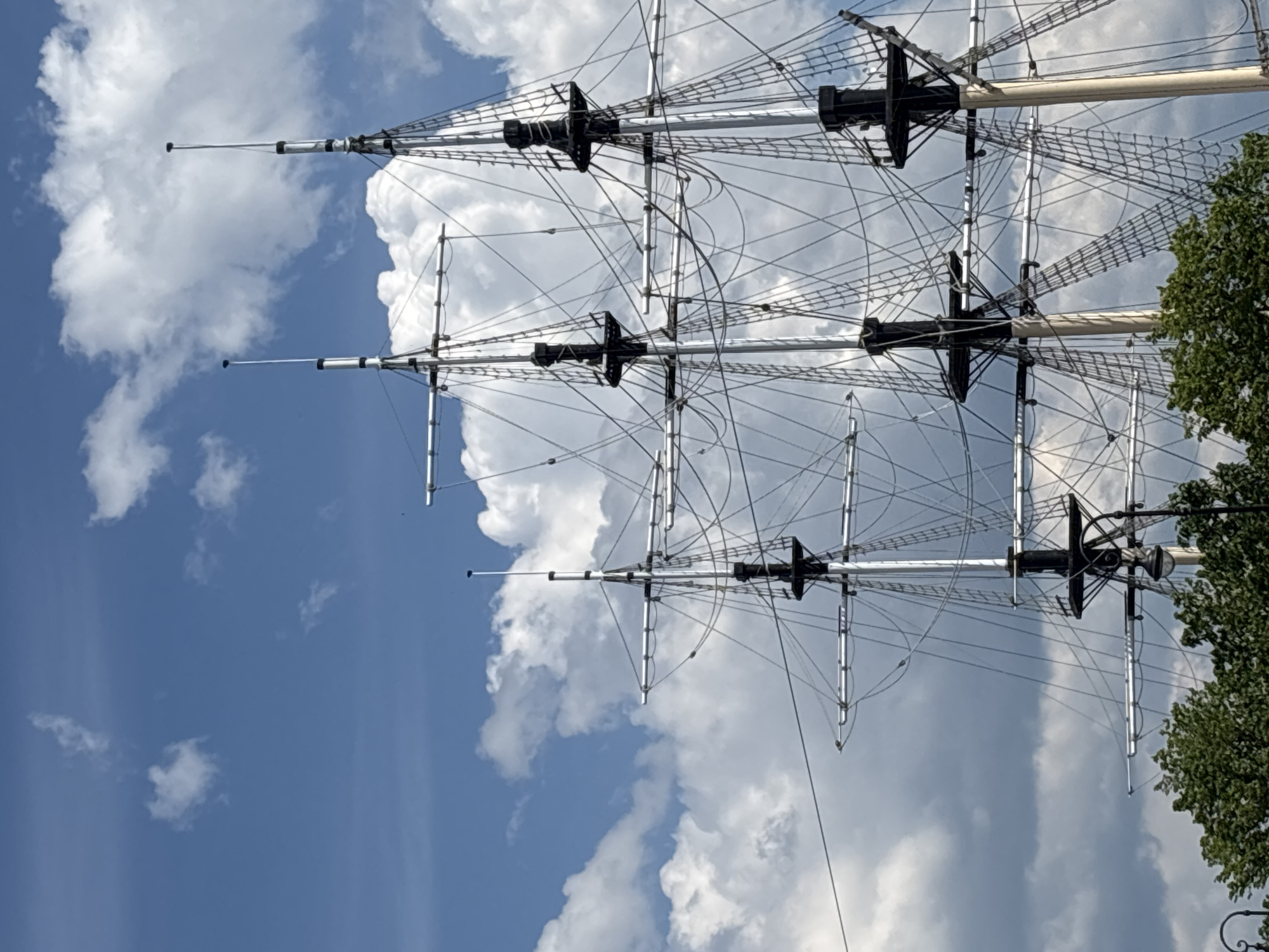 The image presents a detailed, upward-angled view of the upper mast and rigging structures of a large, square-rigged sailing vessel, set against a bright blue sky. Three distinct masts are prominently featured, extending vertically from the lower edge of the frame towards the top.
Each mast appears to be segmented, with lighter-toned main spars and darker, cylindrical sections forming mast tops and platforms. Multiple horizontal spars, known as yards, are affixed perpendicularly to each mast, characteristic of a traditional square rig. A dense and intricate network of black lines, representing shrouds, stays, and various running rigging, crisscrosses between the masts and yards. Ladder-like structures, identified as ratlines, are clearly visible ascending the lower sections of the masts, particularly on the rightmost mast, designed for crew ascent.
The background consists of a vibrant blue sky, partially obscured by numerous bright white cumulus clouds of varying sizes and densities, suggesting fair weather and ample daylight. The very bottom edge of the image reveals the dense green foliage of deciduous trees, partially obscuring the bases of the masts and indicating land or an embankment nearby.
No human figures are visible, nor are any specific architectural landmarks that would definitively pinpoint the exact location within the city. However, given the instruction to include the location in Saint Petersburg, Russia, this scene most likely depicts a tall ship moored along one of the city's prominent waterways, such as the Neva River, or within a historic port area like those near the Admiralty or Vasilyevsky Island, where such large sailing vessels are commonly seen or showcased.