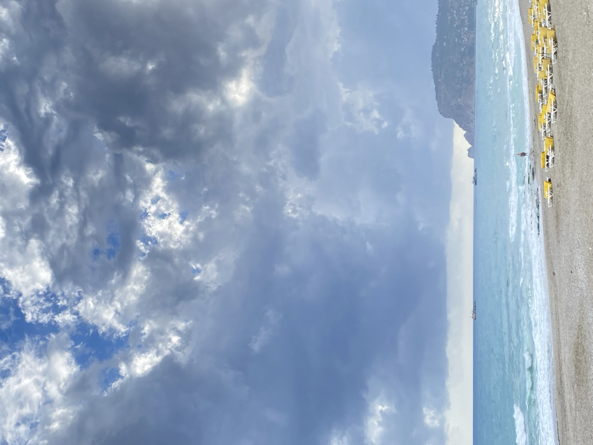 The image shows a beach scene in Alanya, Turkey, featuring a coastline with a mountain backdrop. The sky is overcast with heavy, textured clouds. The sea is light blue, with small waves washing onto the sandy shore. On the right side of the frame, there are rows of yellow beach chairs and some figures, possibly beachgoers. A vessel is visible on the water. The general environment suggests a cloudy day at the beach.