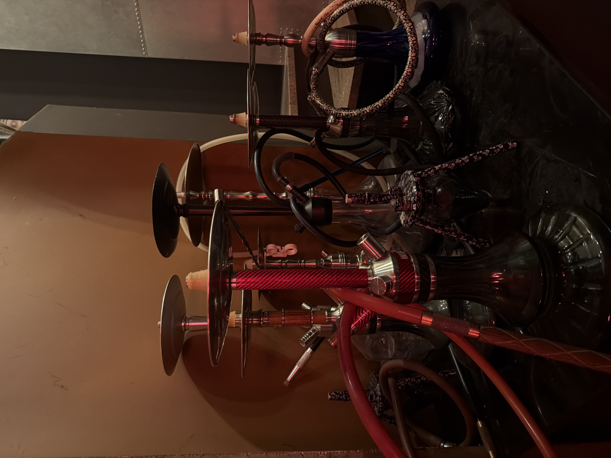 The image depicts a collection of hookahs, likely inside a bar or lounge, potentially in the city of Bar, Montenegro. The hookahs are arranged vertically, each featuring a base, stem, tray, and hose. The bases vary in color, including blue, clear, and black. The stems exhibit different designs and materials, such as carbon fiber and metal. Hoses are red, black, and patterned. The backdrop consists of a textured wall and a dark surface reflecting the hookahs.
