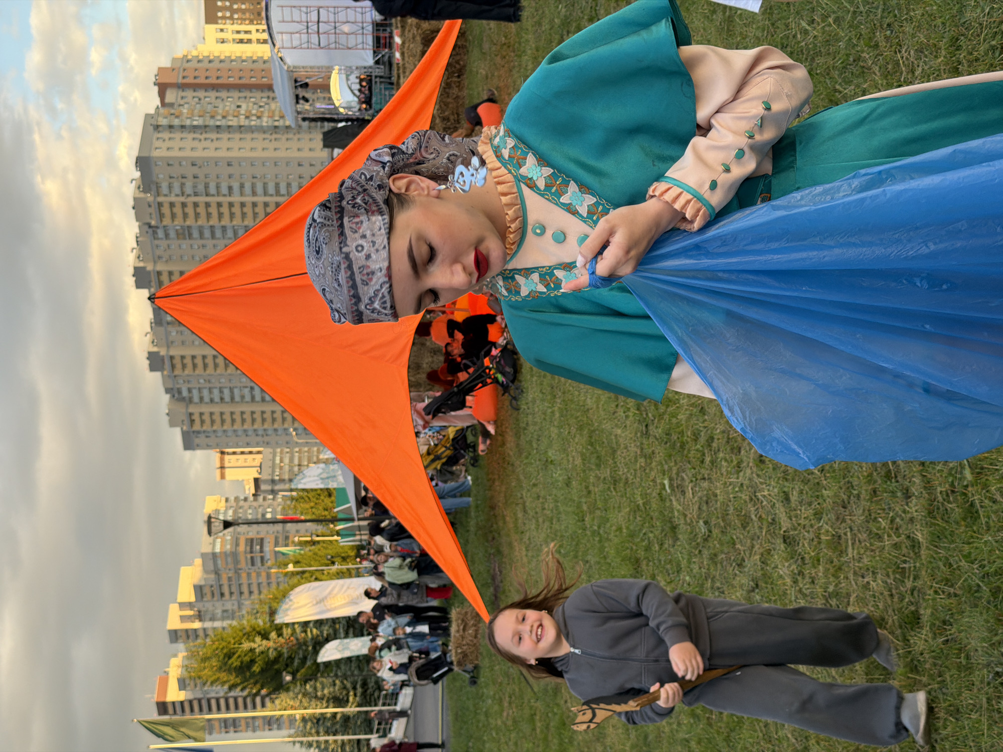 In an outdoor grassy area, a young girl with long brown hair, wearing a grey hooded sweatshirt and grey pants, smiles as she holds a wooden stick. To her right, a young woman in a teal and beige traditional outfit with a patterned headscarf and large blue earrings is holding a blue plastic bag. In the background, people are gathered near a large orange tent, with flags and apartment buildings visible under a cloudy sky.