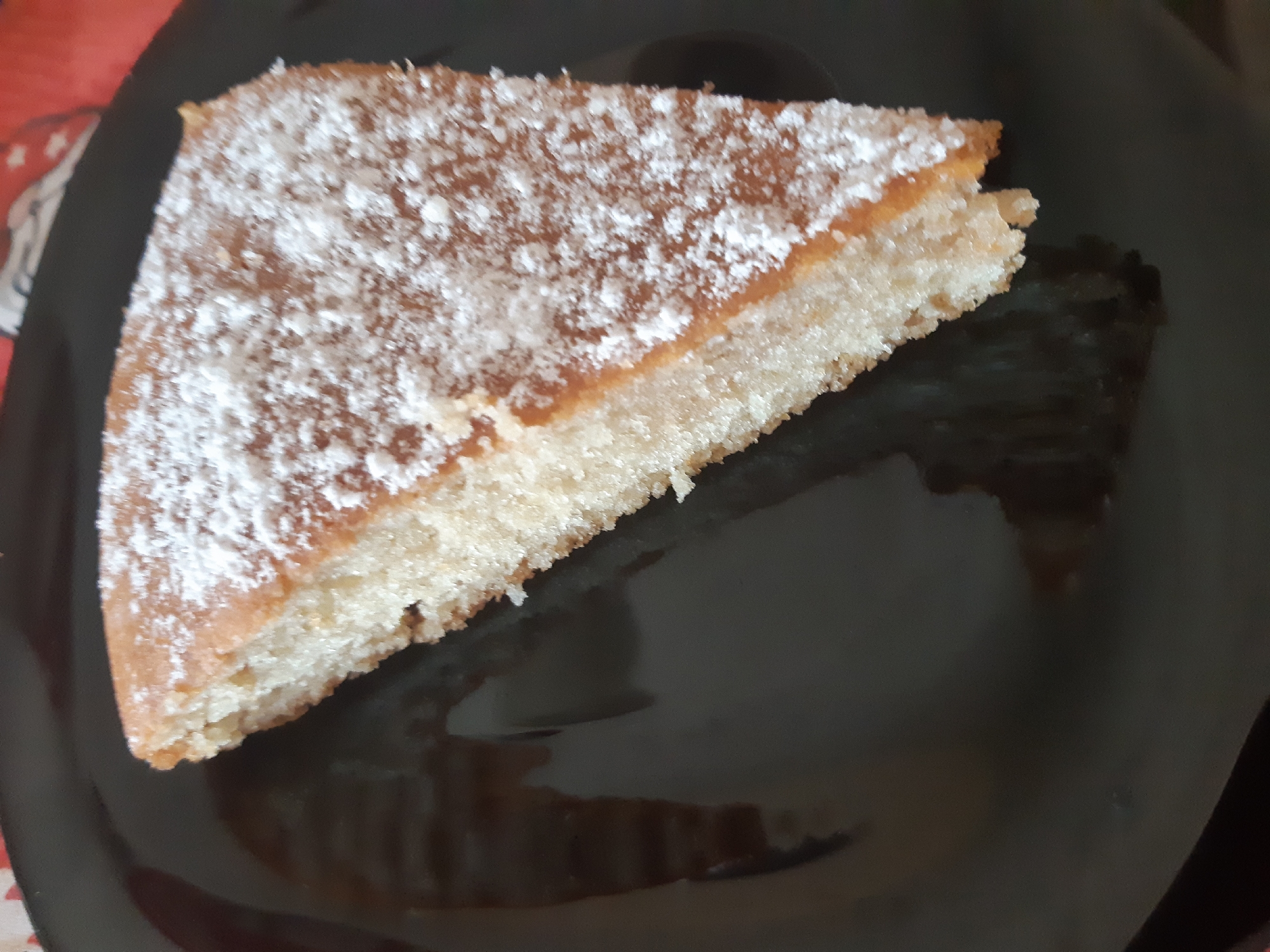 A triangular slice of cake, dusted with powdered sugar, rests on a black plate. The cake has a light-colored base and a slightly darker upper layer. The plate appears glossy, reflecting light. There is no indication of the city of Plovdiv, Bulgaria, or its presence in the image.