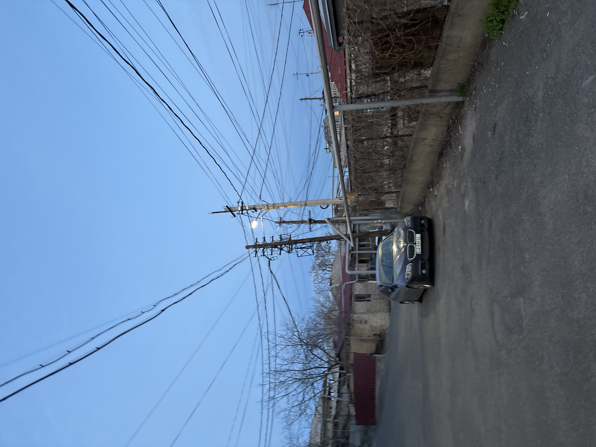 The image captures a street scene in Yerevan, Armenia. The sky is pale blue. Overhead, multiple power lines stretch across the frame. On the right side of the image, a narrow asphalt road is visible. A dark-colored car is parked on the side of the road. Adjacent to the road is a concrete barrier covered with vegetation. Behind the barrier, a building with a red roof is seen, featuring a metal pipe running along the wall. Electrical poles are erected along the street. Sparse trees with bare branches are visible. The angle of the shot is slightly tilted.