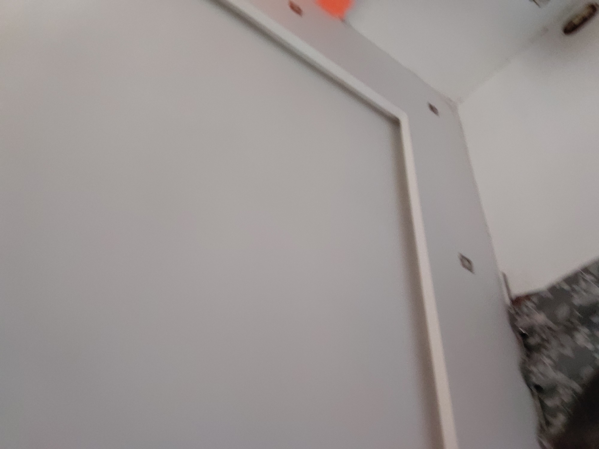 The image presents an interior view, characterized by a soft focus. A large, plain, light-gray surface, possibly a wall or panel, dominates the left and central portions of the frame. This surface is outlined on its right by a prominent white trim or frame.
Above and to the right of this main surface, a white ceiling is visible. An orange object with an irregular shape is attached to the ceiling in the upper left corner. Further to the right on the ceiling, a blurry, dark, circular object, which might be a light fixture or vent, can be seen.
To the right of the white trim, on an adjacent wall or structure, a small, dark, and somewhat diamond-shaped object is visible. On the extreme right edge of the image, a section of a blurry, patterned material, appearing to be a mix of gray and dark tones, is partially visible, suggesting a fabric with a camouflage or abstract design.
The scene depicts a static indoor environment. No specific activity or event is taking place. The time of day and external weather conditions cannot be determined. There is no visible text in the image. The overall impression is that of a functional, possibly industrial or utility space.