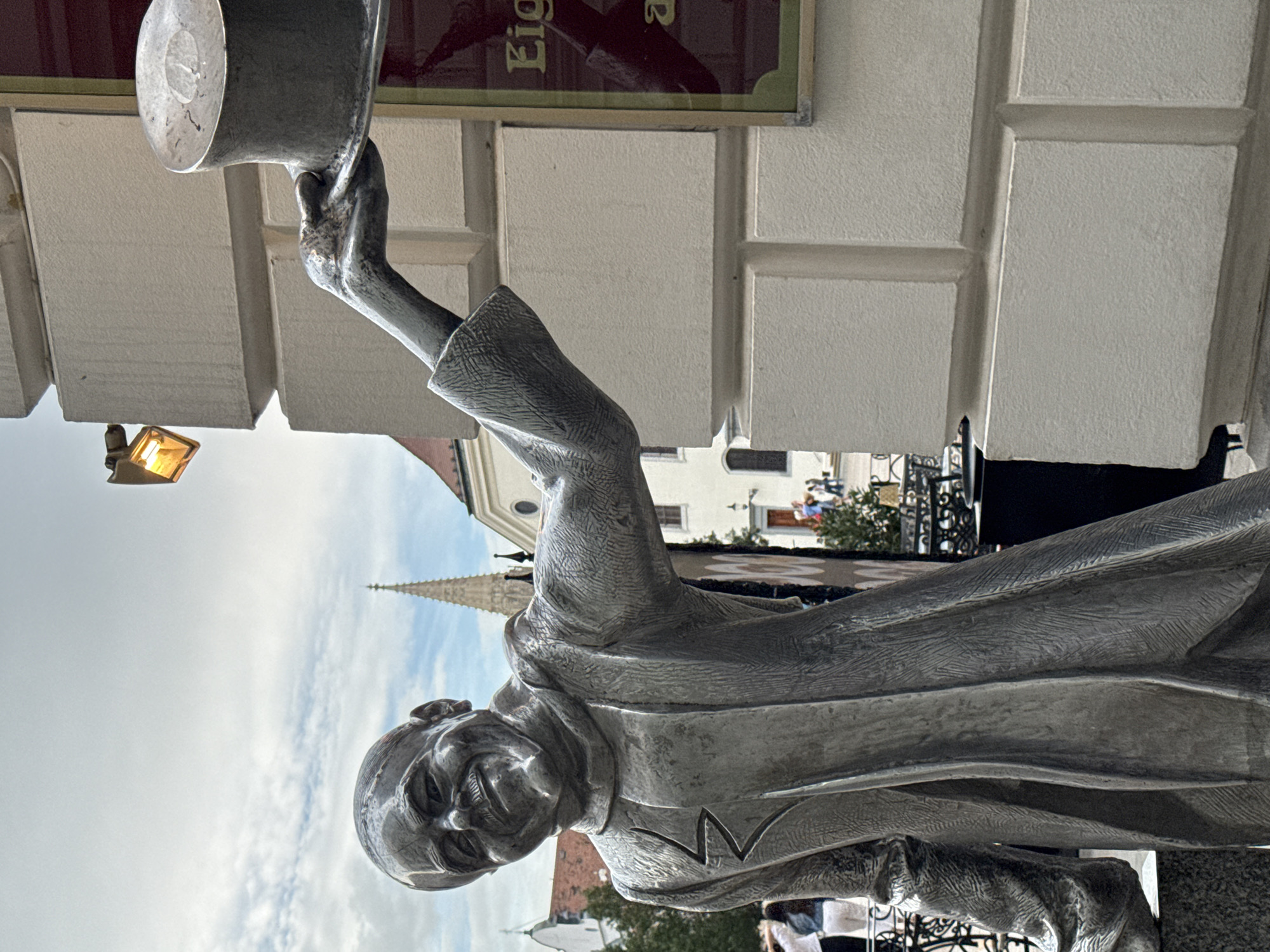 The image shows a close-up of a silver statue of a man wearing a coat and holding a top hat in his raised right hand. The statue is positioned next to a light-colored stone building with a sign on it. The statue is life-sized and appears to be located outdoors, possibly on a street or in a public square. The background shows a cloudy sky, a church spire, and other buildings. People can be seen in the background, indicating a public space. The statue is likely a monument to a notable figure. This statue is located in Bratislava, Slovakia.