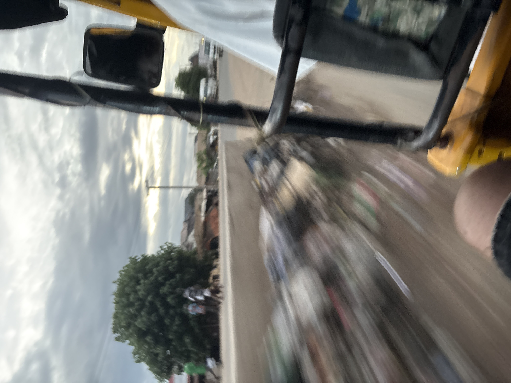 This image captures a blurry, motion-filled view from inside a yellow motorized tricycle, likely a "keke-napep" or similar, traveling through Onitsha, Nigeria. The foreground is dominated by the tricycle's yellow frame and black handlebars, with a portion of a person's leg visible on the lower left. To the left of the tricycle, a wall of accumulated refuse and debris is visible, blurred due to motion. Beyond the wall, a street scene unfolds with a large, green tree, several people walking, and buildings in the background. The sky above is overcast with dramatic clouds. Further down the road, more vehicles, including a white truck, are visible. The overall impression is one of a busy urban environment in motion.
