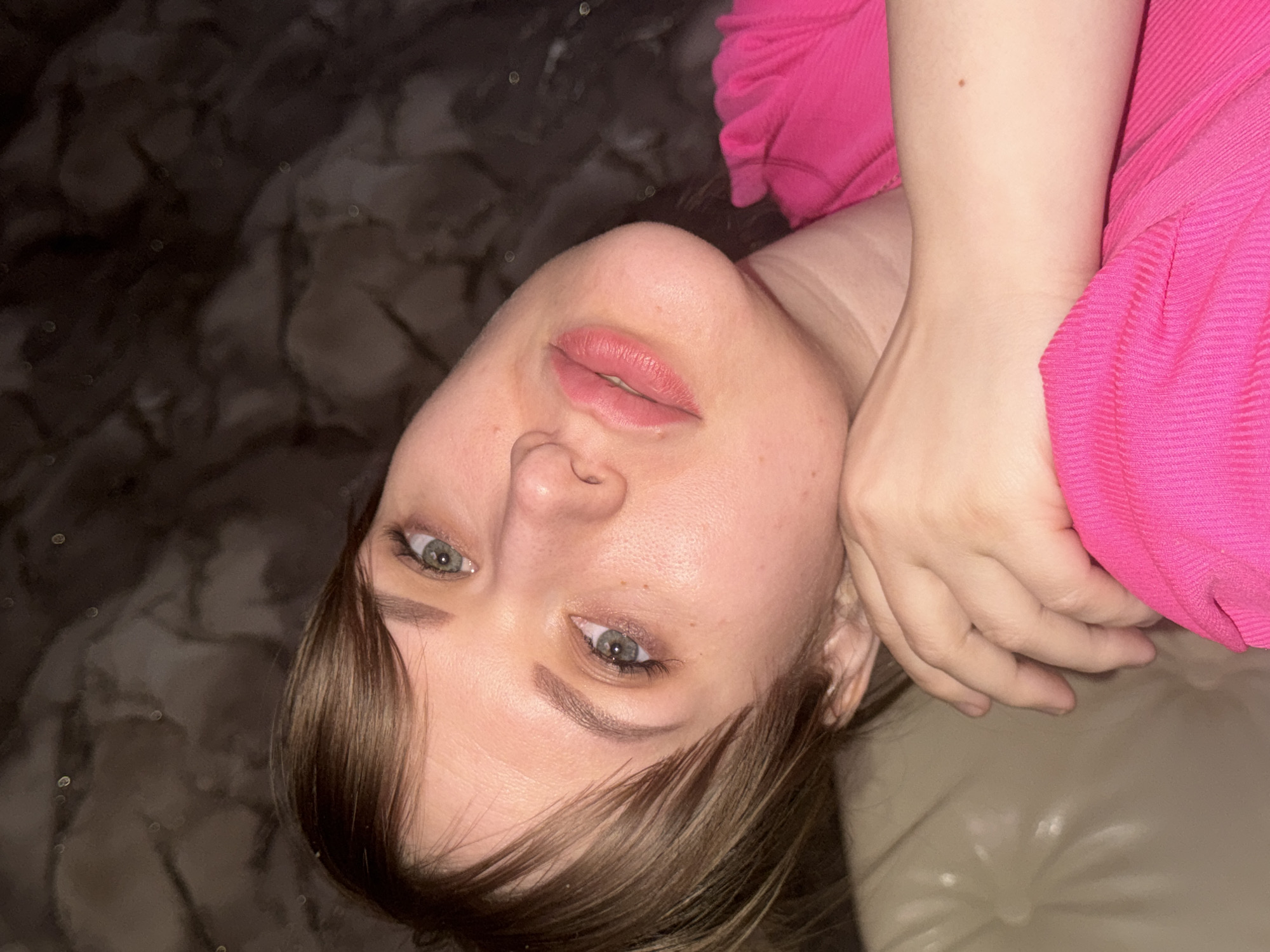 This close-up portrait shows a young woman in an indoor setting, possibly in Chaikovsky, Russia. She is depicted from the chest up, with her head rotated approximately 90 degrees counter-clockwise, looking directly towards the viewer with a calm expression. She has fair skin, blue-green eyes, and light brown hair with subtle lighter highlights, which partially covers her forehead and her right eye. Her lips are soft pink and slightly parted.
She is wearing a bright pink, textured top or dress. Her left arm is bent, with her hand resting near her head. She appears to be lying on a light-colored surface, possibly a sofa or bed, which is visible on the right side of the frame. The background on the left is dark and mottled, possibly a patterned fabric or wall, featuring small, blurred circular lights (bokeh), indicating a low-light environment, likely evening or nighttime. The overall scene conveys a relaxed and contemplative mood. No text is visible in the image.