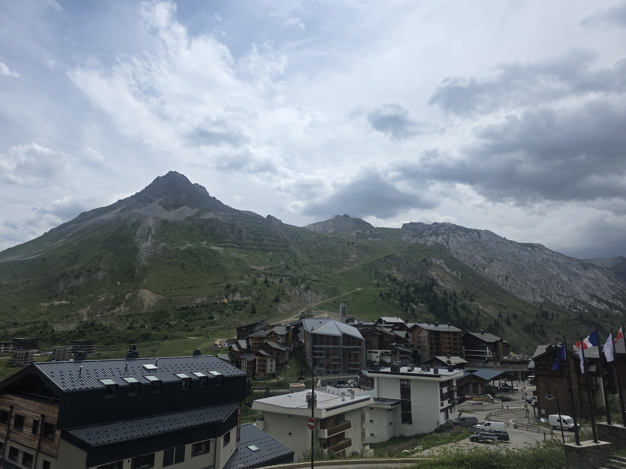 The image depicts a panoramic view of the ski resort town of Tignes, France, nestled at the foot of a vast mountain range under a cloudy sky. The foreground features several multi-story buildings with dark, sloped roofs, some exhibiting wooden facades and balconies. In the middle ground, a cluster of chalet-style buildings with pitched roofs and balconies forms the heart of the town, characterized by their warm wood tones and stone accents. A few modern buildings with contemporary architectural designs are interspersed among the traditional structures. Roads wind through the town, with a few cars and vans parked or driving. The background is dominated by the imposing mountain landscape, with rugged, rocky peaks and steep, green slopes dotted with sparse vegetation and some trees. The sky is filled with a dramatic display of grey and white clouds, suggesting an overcast or approaching weather system. Flags, including the French tricolor, are visible on poles in the right foreground. The scene conveys the alpine environment of Tignes, a popular destination for winter sports and summer mountain activities.