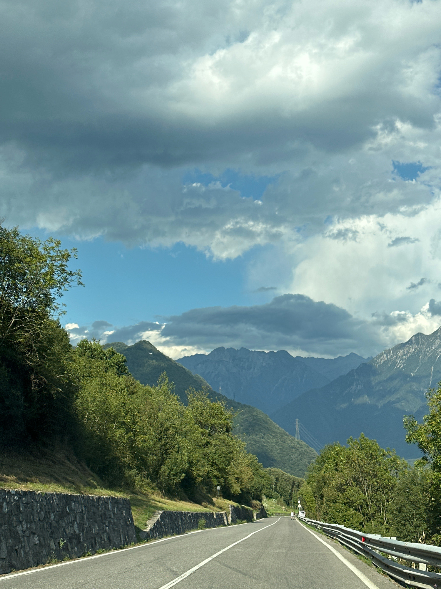 The image depicts a road winding through a mountainous landscape, possibly near Ossimo Inferiore, Italy. The road, paved with asphalt and marked with white lines, curves to the right in the foreground. On the left side of the road, a stone retaining wall supports a grassy embankment covered in green vegetation. Trees and dense foliage line the upper edge of the embankment. In the distance, a series of mountain ranges are visible, their peaks shrouded in a hazy blue. The sky above is dramatic, filled with a mix of dark, heavy clouds and patches of bright blue sky, with some lighter, more scattered clouds. A transmission tower or pylon can be seen on one of the distant slopes. The overall impression is of a scenic drive through a rural, mountainous region under a partly cloudy sky. There are no identifiable people or specific structures that would pinpoint the exact location beyond the general mountainous terrain.