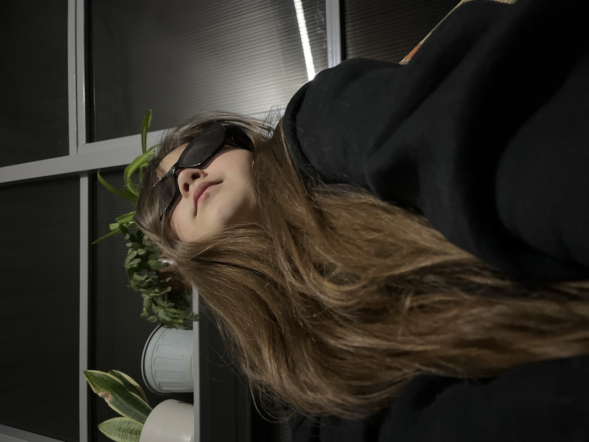 A person with long brown hair is wearing sunglasses and a black hooded sweatshirt. The person is indoors, with a window behind them. Potted plants are visible near the window. There is no visible information to indicate the city of Novopokrovskaya, Russia.