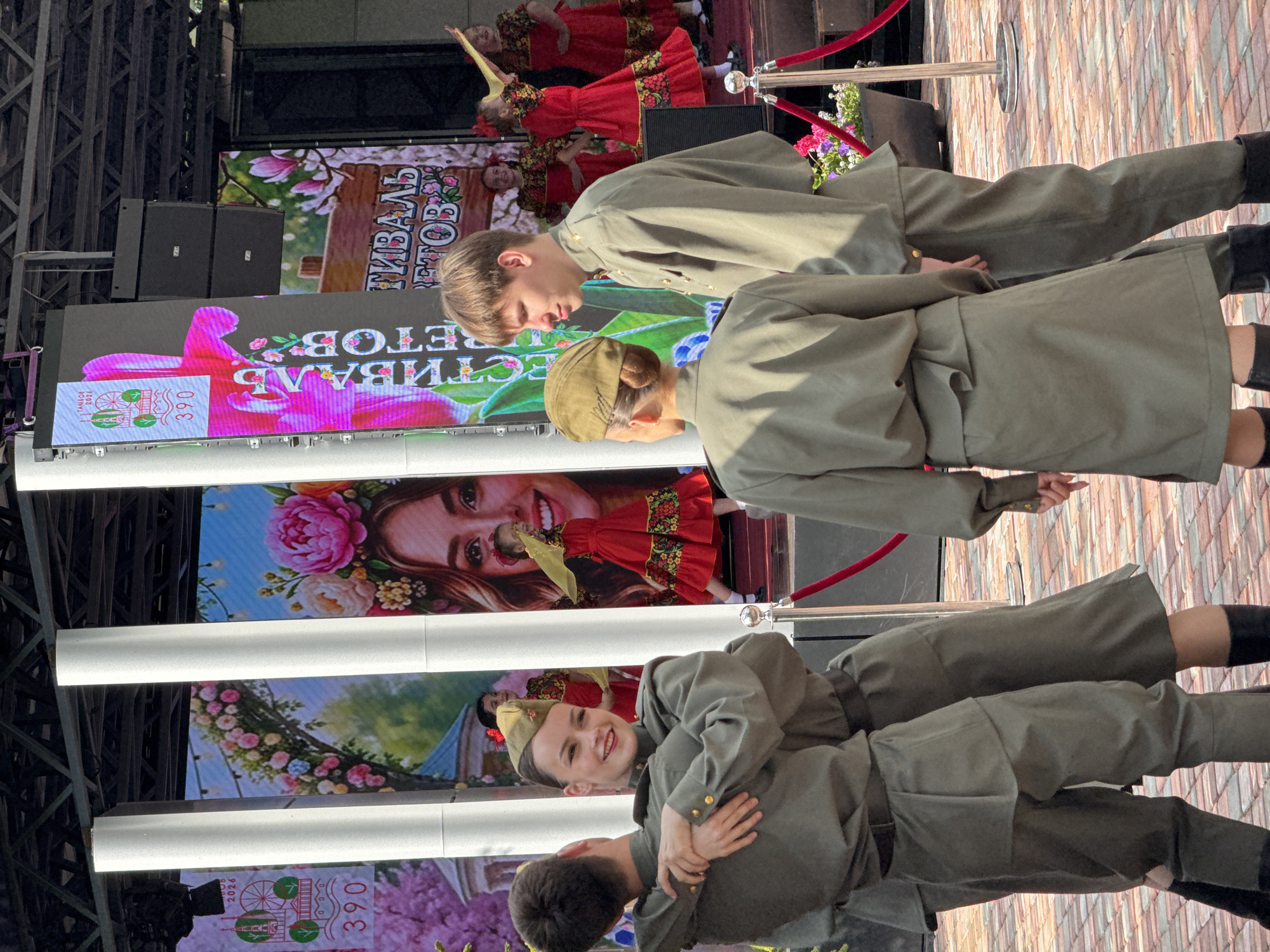 Here is a detailed description of the image:
The image shows three individuals dressed in military-style uniforms, likely performing on a stage. The backdrop features large screens displaying various images, including flowers, portraits of people, and promotional material. The performers appear to be engaged in a choreographed dance or performance, with two of them in the foreground and one further back. The overall setting suggests an outdoor event or festival, possibly in the city of Unknown, Unknown. The stage is set up with red ropes as barriers, and the ground appears to be paved.