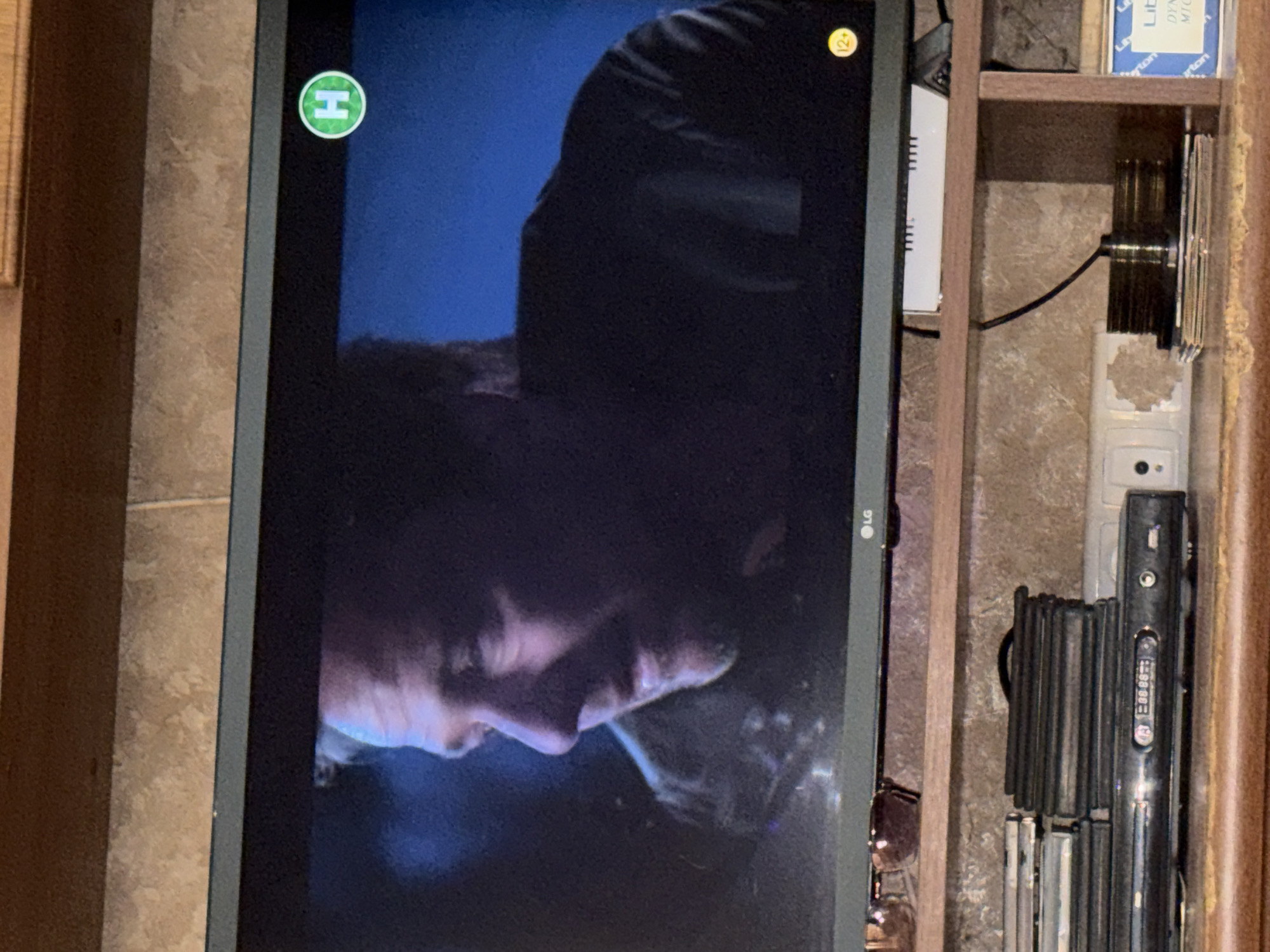 A medium shot captures a television screen displaying a man's face in profile against a dark blue background. The man appears to be middle-aged, with light-colored hair, and his facial features are partially obscured by shadows. A green circular logo with a white "H" is visible in the upper right corner of the screen. In the lower right corner of the screen, a yellow circular icon with "12+" is present. The bottom edge of the television screen shows the "LG" logo. The television is situated on a wooden shelf, below which are more shelves containing various items. These include a stack of what appear to be DVDs or similar media, a DVD player with a digital display showing "08:88:88", and a stack of round objects, possibly discs, on a metal spindle. To the far right, a blue framed object with text is partially visible. A pair of sunglasses rests on a shelf above the television. The background appears to be a tiled wall. The scene is dimly lit, suggesting an indoor setting, likely a home entertainment area.