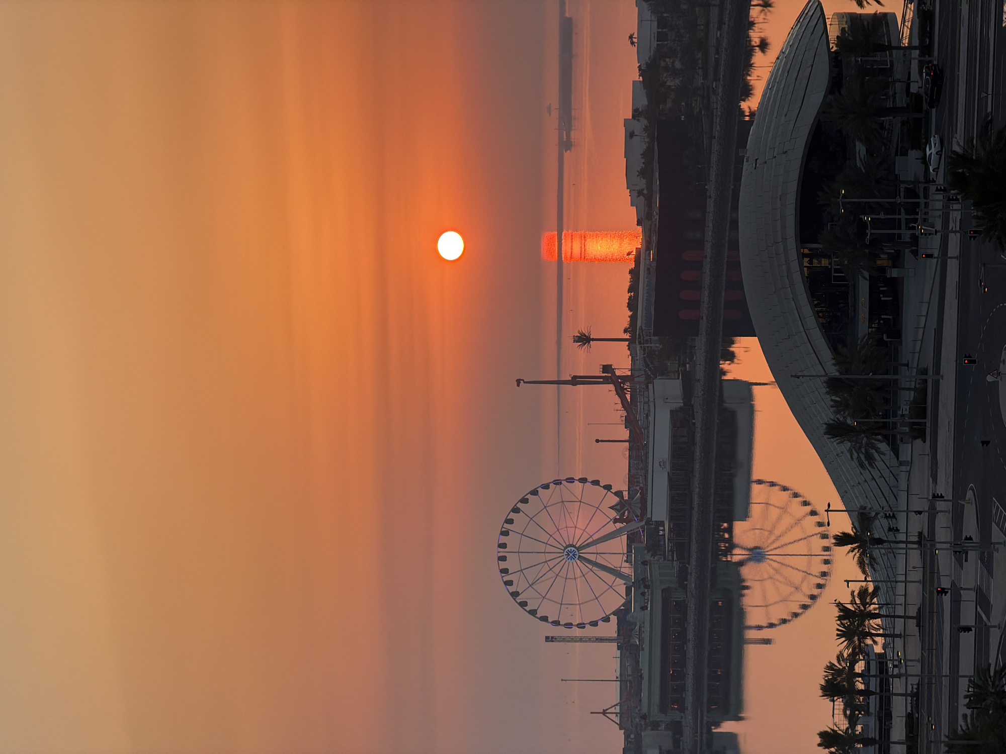 A wide shot captures a vibrant sunset over a cityscape. The sky is painted in gradients of orange and red, with the sun appearing as a bright, distinct orb. The sun's reflection shimmers on the water below, creating a luminous vertical streak. In the mid-ground, a large Ferris wheel stands illuminated with blue lights, and a collection of buildings, some with amusement park structures, line the waterfront. Palm trees are scattered throughout the scene. In the foreground, a modern building with a distinctive curved roof is visible, along with a street with traffic lights and cars. The water acts as a mirror, reflecting the Ferris wheel and the surrounding structures. The overall scene is one of a coastal city at dusk.
