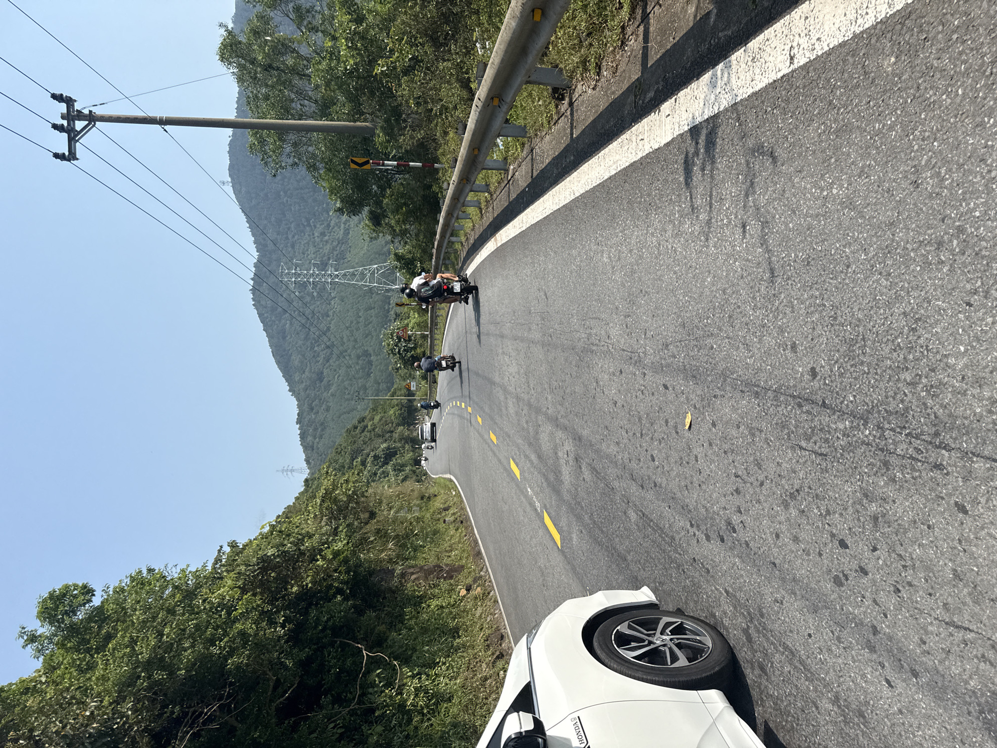 The image shows a winding road in Đà Nẵng, Vietnam, bordered by lush green trees and mountains in the background. A white car is partially visible in the foreground on the right side of the road. Several motorcycles are traveling along the road. Overhead, power lines stretch across the scene. A metal guardrail runs alongside the road on one side, delineated by a solid white line on the road surface.