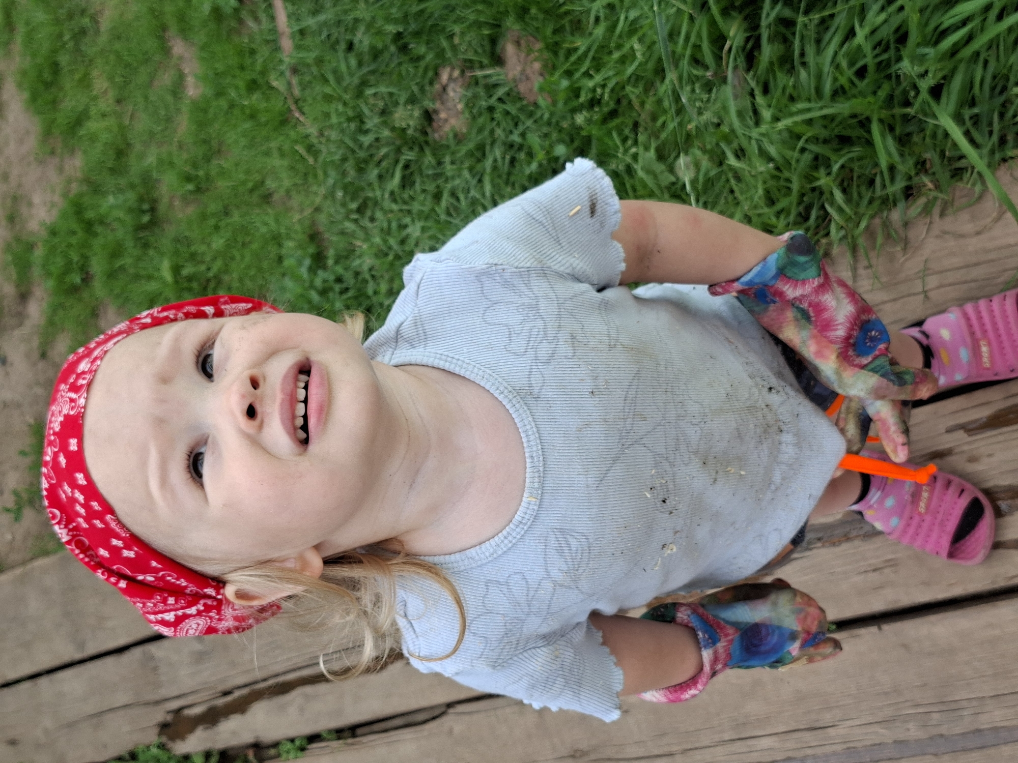The image shows a young child, possibly a toddler, standing on a wooden walkway next to a grassy area. The child is wearing a red bandana on their head, a light blue t-shirt that appears to be dirty, gardening gloves with a floral pattern, pink sandals, and pink socks. The child's face is looking upwards with a pained or questioning expression. The environment appears to be outdoors, with green grass and a wooden surface. The image does not provide any specific information or context that would link it to the city of Karsha, Russia.