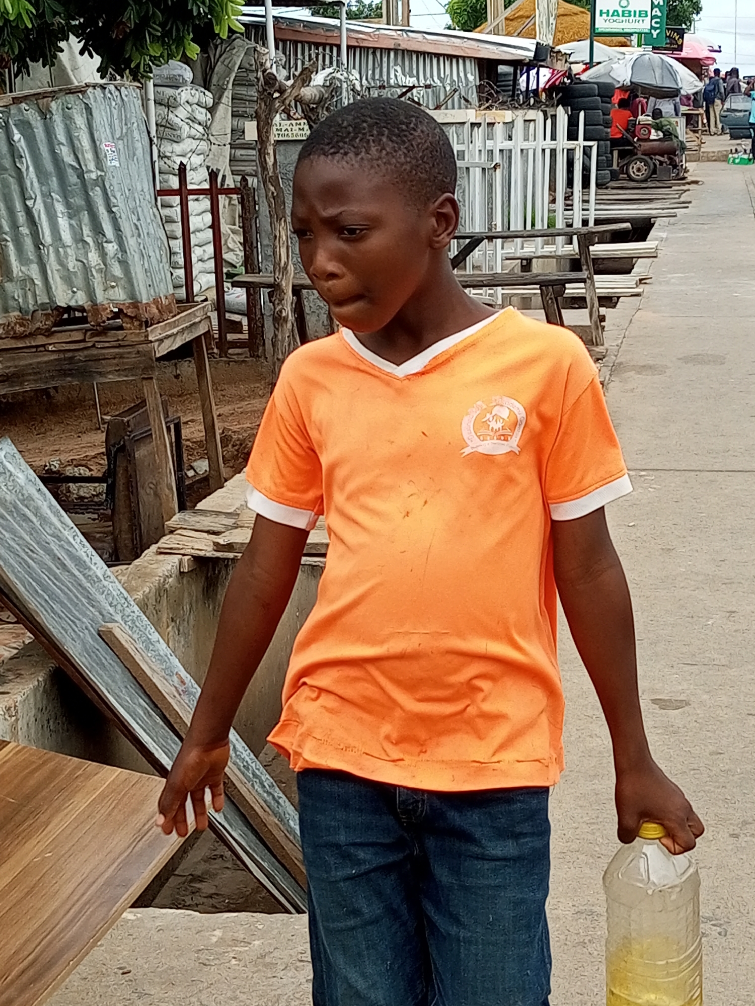 This media file captures a young boy in an outdoor, bustling market setting in Tempure, Nigeria. The boy, of African descent, is wearing an orange V-neck t-shirt with a white trim and dark blue jeans. He is holding a large, clear plastic bottle filled with a yellow liquid, likely some form of edible oil or drink, in his right hand. His facial expression appears thoughtful or concerned.
The background reveals a vibrant street scene typical of a Nigerian market. Stalls are set up with various goods, including what appear to be stacked tires, corrugated metal structures, wooden benches, and possibly some food items. In the distance, there are more stalls with awnings and people milling about, suggesting the market is active. The ground is a paved pathway, and there are trees and vegetation visible, hinting at a warm, possibly humid climate.
A prominent sign in the background reads "HABIB YOGHURT", indicating a food vendor. Other signs are partially visible and difficult to decipher. The overall lighting suggests it is daytime, likely late morning or afternoon, with natural sunlight illuminating the scene. The atmosphere appears lively and somewhat chaotic, typical of a busy market. The boy's focus seems to be directed forward, and his posture suggests he is on the move, possibly carrying the bottle to a destination. The image provides a snapshot of everyday life in a Nigerian market, highlighting the commerce and movement within such a setting.
