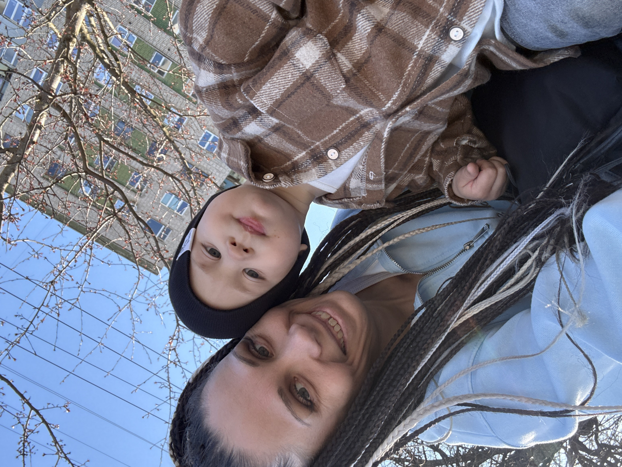 An upward-facing selfie captures an adult female and a young child outdoors on a bright, sunny day, with the image appearing rotated. The adult, positioned in the foreground, smiles at the camera, showcasing long, dark and light braided hair and wearing a light blue zippered top. To her left, a young child with fair skin and light eyes looks towards the camera, wearing a black beanie and a brown and white plaid shirt over a white top. The child's small hand is visible near the adult.
The background features a clear blue sky. Bare tree branches with visible buds stretch across the frame, indicating early spring. Behind the trees, a tall, multi-story residential building with numerous windows and a light-colored facade suggests an urban or suburban setting. The bright daylight implies the photo was taken during the day. Both subjects appear relaxed. No text is visible in the image.