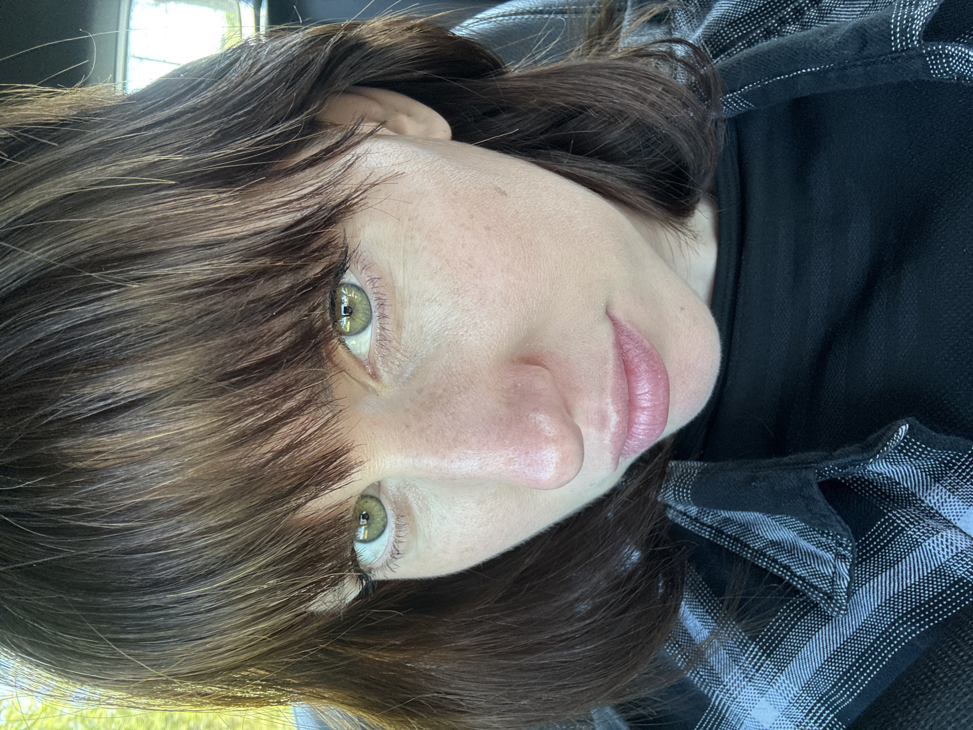 A close-up headshot features a woman with medium-length, dark brown hair styled with bangs. Her skin is fair with freckles. The eyes are light green or hazel in color. The woman is wearing a black shirt and a black and white plaid collared shirt over it. The background is out of focus but suggests an outdoor setting with some light and greenery, though the specific location in Nettuno, Italy, is not discernible from this image.