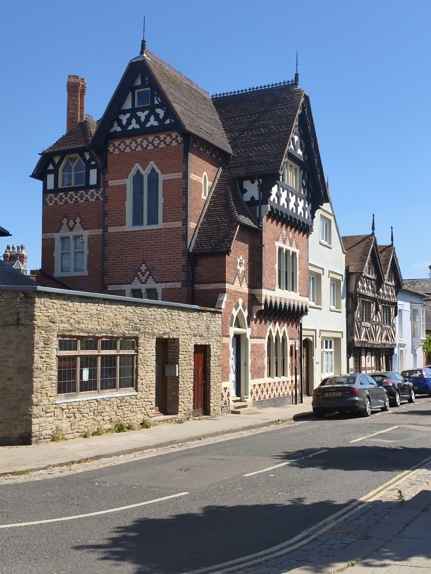 This is a high-angle, medium-shot photograph of a row of buildings on a street in Ludlow, United Kingdom, on a bright, clear day with a blue sky. The buildings are varied in architectural style and age, featuring red brick, stone, and timber-framed facades.
The most prominent building on the left is a tall, red brick structure with decorative white and black timber framing. It has a steep, tiled roof with ornate gables and a chimney. To its right, a white building with a white and timber-framed facade follows, with a pitched tiled roof and dormer windows. Further to the right, another building with a black and white timber-framed facade is visible, characterized by its dark wooden beams and white infill. The buildings on the far right appear to be lighter in color and possibly of a more modern construction.
Parked along the street in front of these buildings are several cars. Closest to the viewer is a dark gray Audi sedan with license plate NG18 BFK. Behind it is a dark-colored car, and further down the street, a blue car is visible. The street itself is paved with asphalt and has white lane markings. A pedestrian sidewalk runs alongside the buildings, constructed with paving stones and some grass growing along the edges. The overall scene depicts a charming, historic streetscape with a mix of residential and possibly commercial properties.