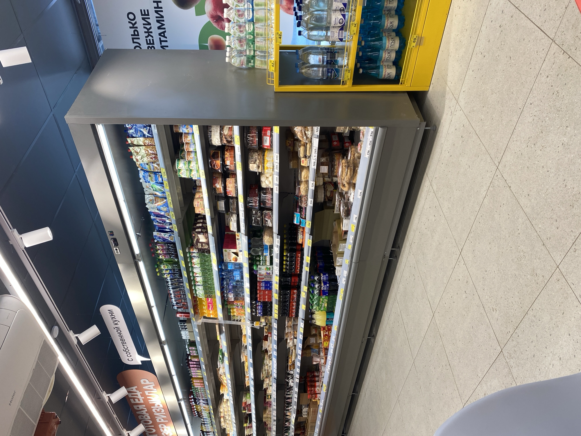 This image shows a refrigerator display in a supermarket, likely located in Kazan, Russia, judging by the Russian writing on the promotional materials. The refrigerator is filled with various food items, including drinks, snacks, and possibly ready-made meals. Above the refrigerator, there are shelves containing bottled water. The setting appears to be a modern, well-lit supermarket with tiled flooring. A sign near the refrigerator section advertises ready-made food with local cuisine.
